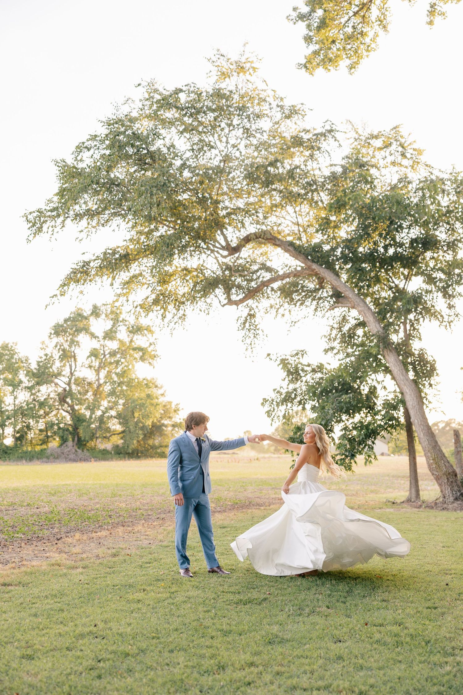 Groom in blue suit spinning bride in white dress under a large tree at golden hour.
