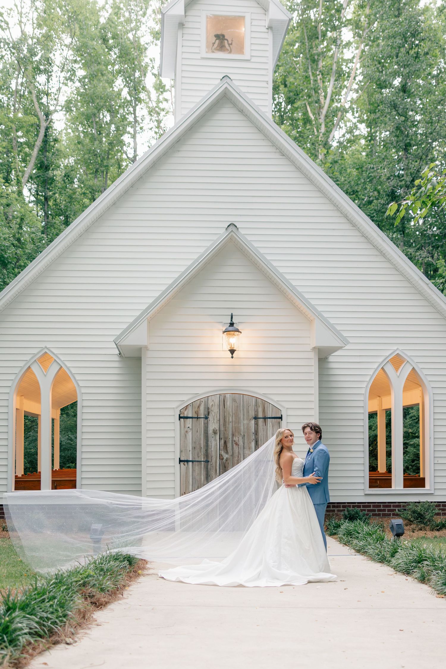 Bride and groom embrace before white chapel with flowing veil billowing.