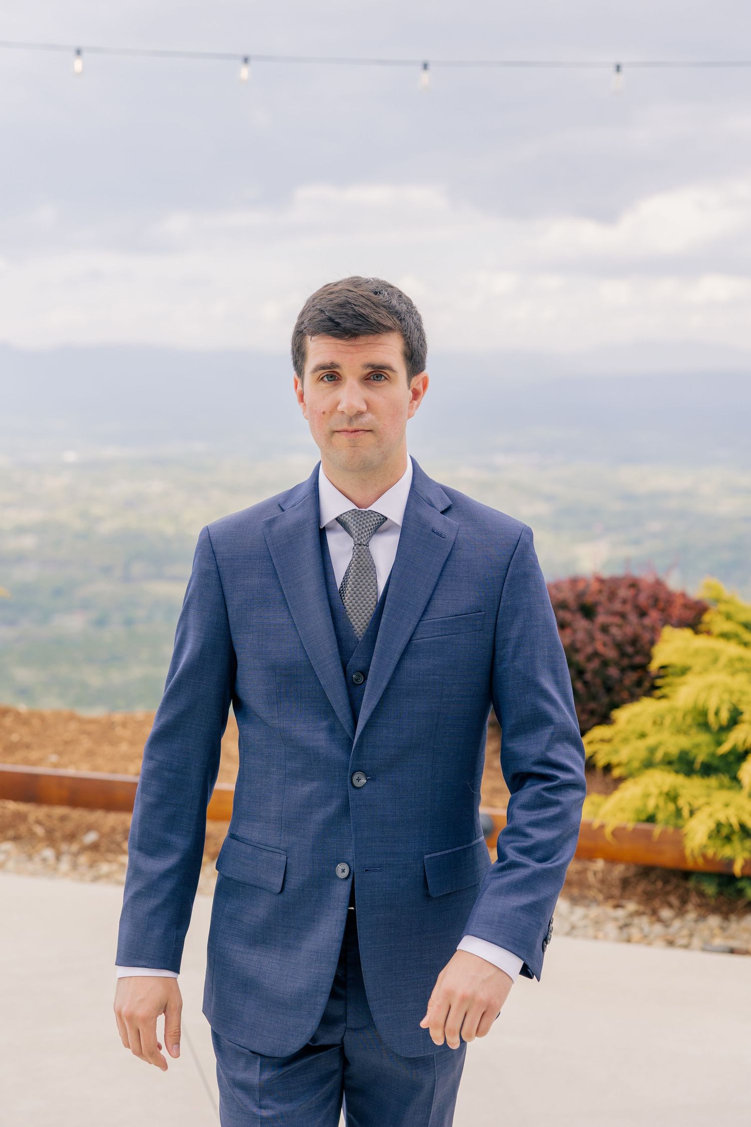 Handsome groom in navy blue three-piece suit standing on outdoor terrace with mountain views.