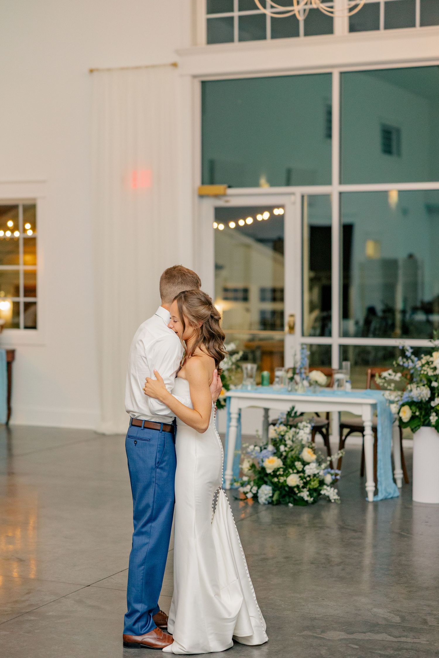 Bride and groom share intimate first dance embrace in elegant white reception venue at night.