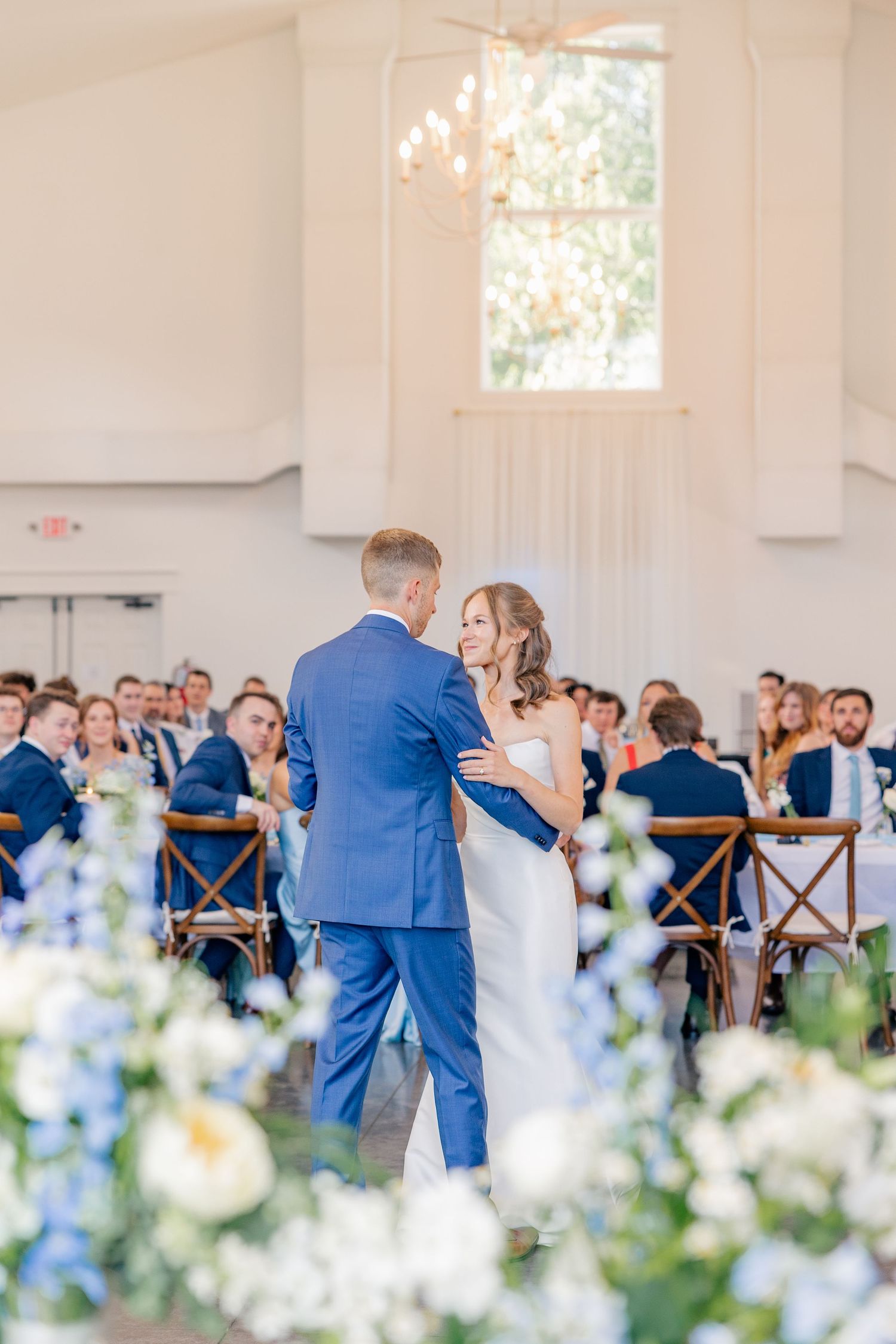 Bride and groom share their first dance in a bright white reception hall with blue and white floral arrangements.