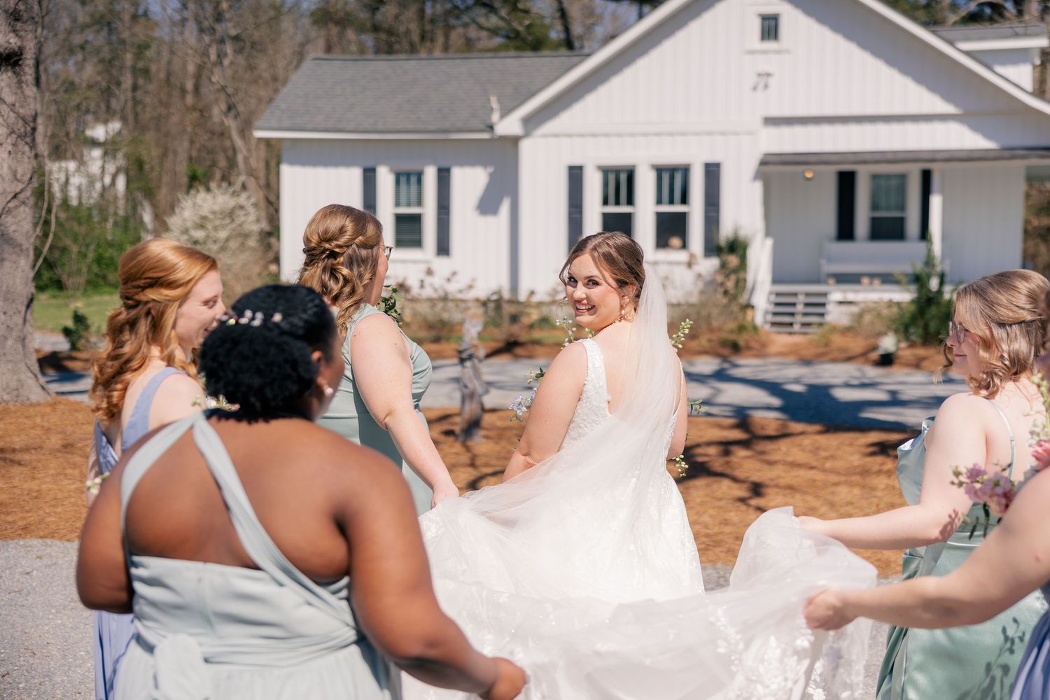 Bridesmaids in pastel dresses laugh and celebrate outdoors on a sunny wedding day.
