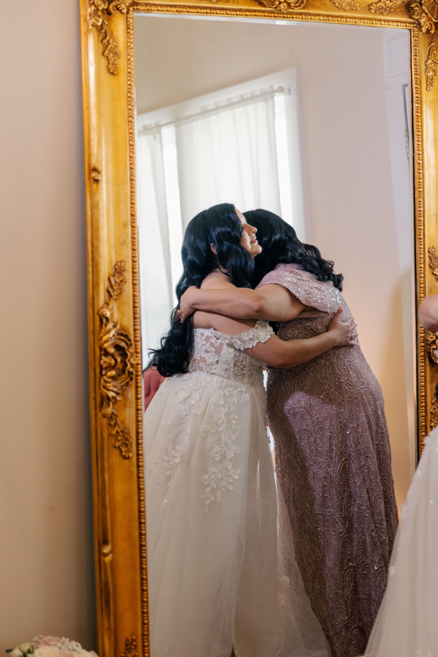 Two figures embrace in an emotional moment before an ornate gold-framed mirror.