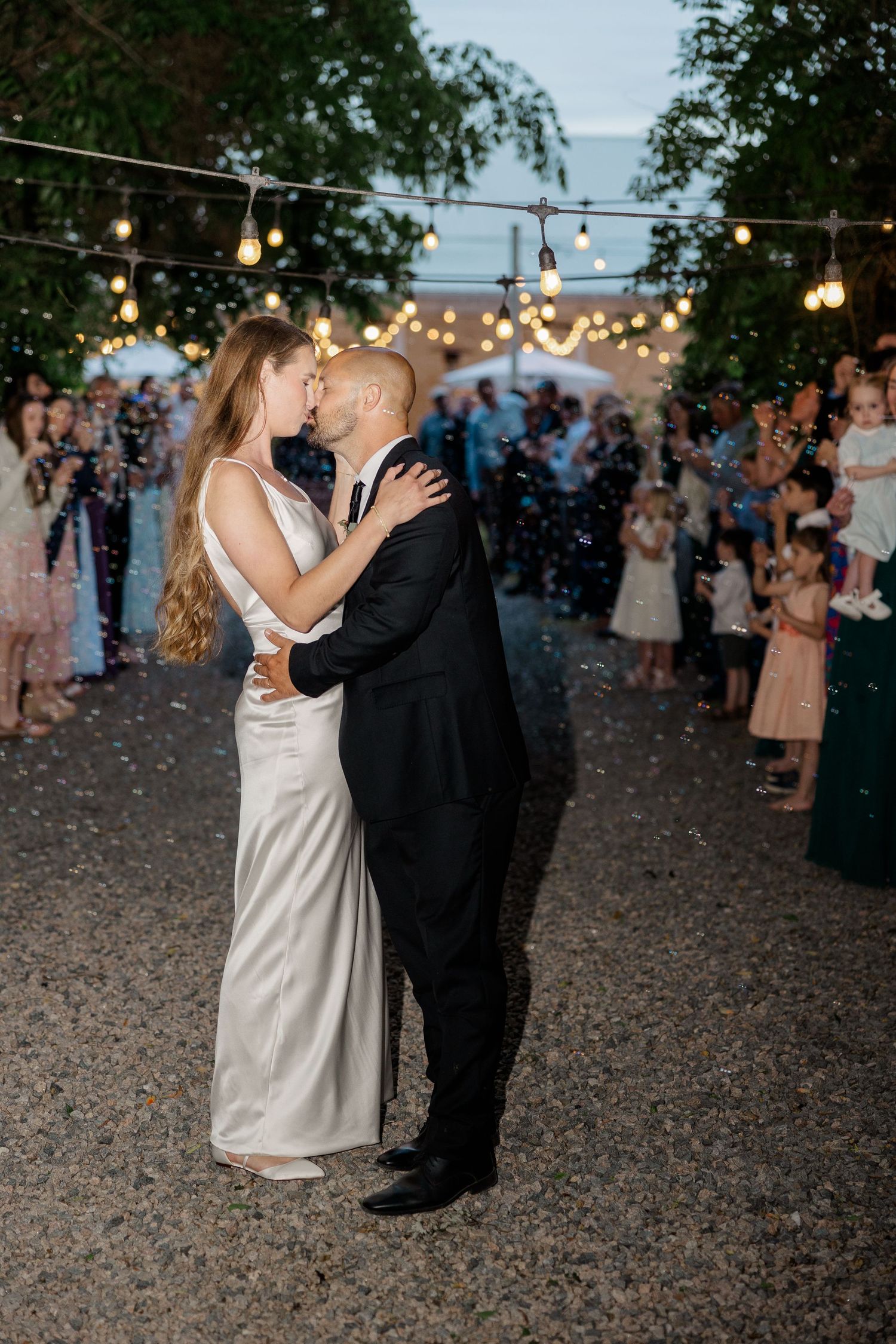 Couple dancing under romantic string lights at dusk during wedding reception celebration.