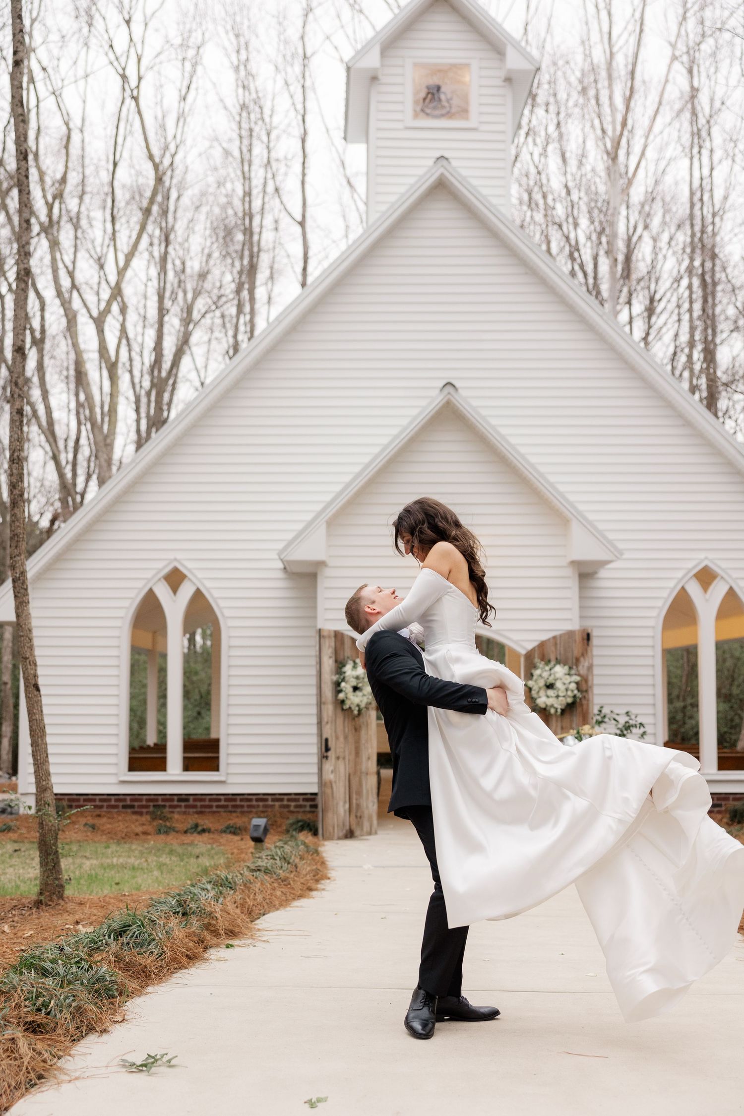 A couple shares a romantic dip pose in front of a white chapel surrounded by trees.