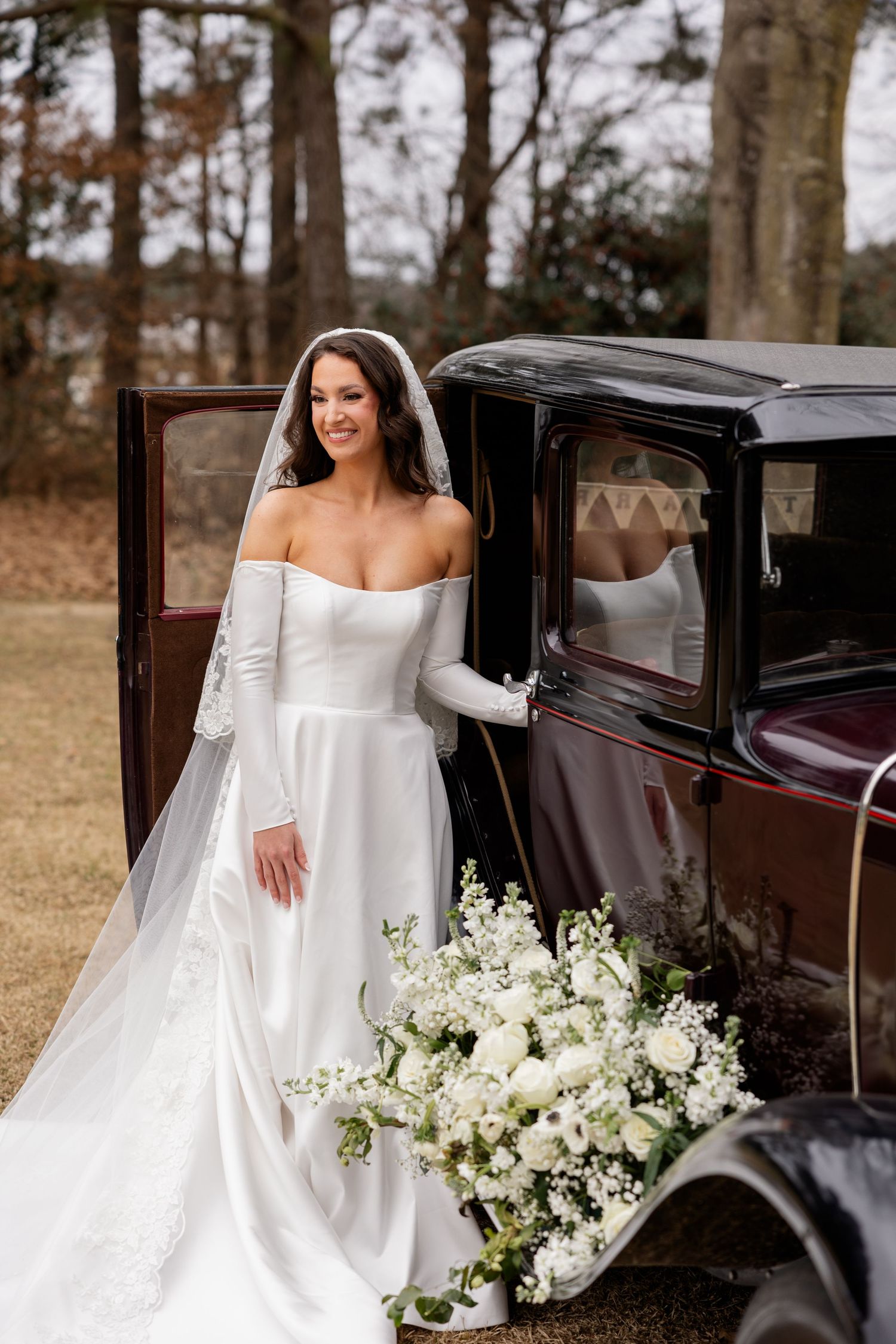 A bride in an elegant white gown poses with a vintage car decorated with white floral arrangements.