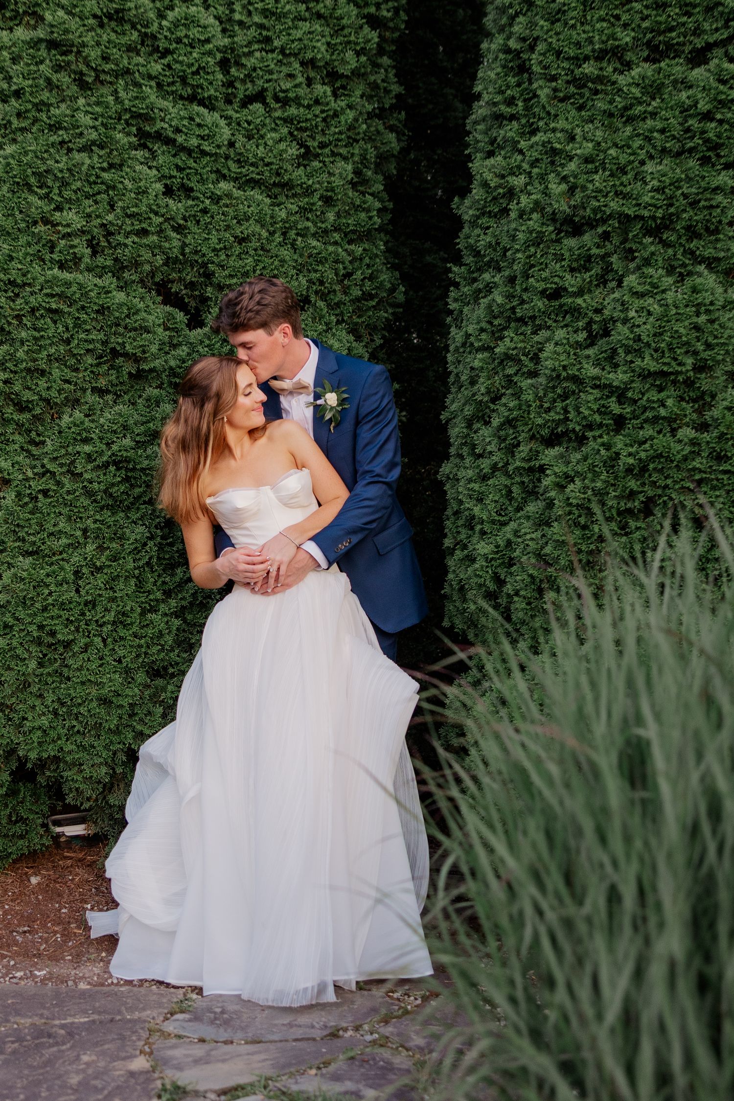 Romantic wedding couple embracing against a backdrop of manicured hedges and ornamental grasses.