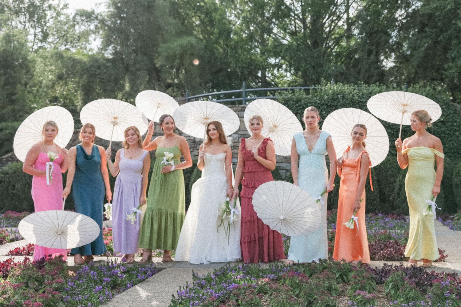 A wedding party in colorful pastel dresses stands in a line outdoors holding white parasols.