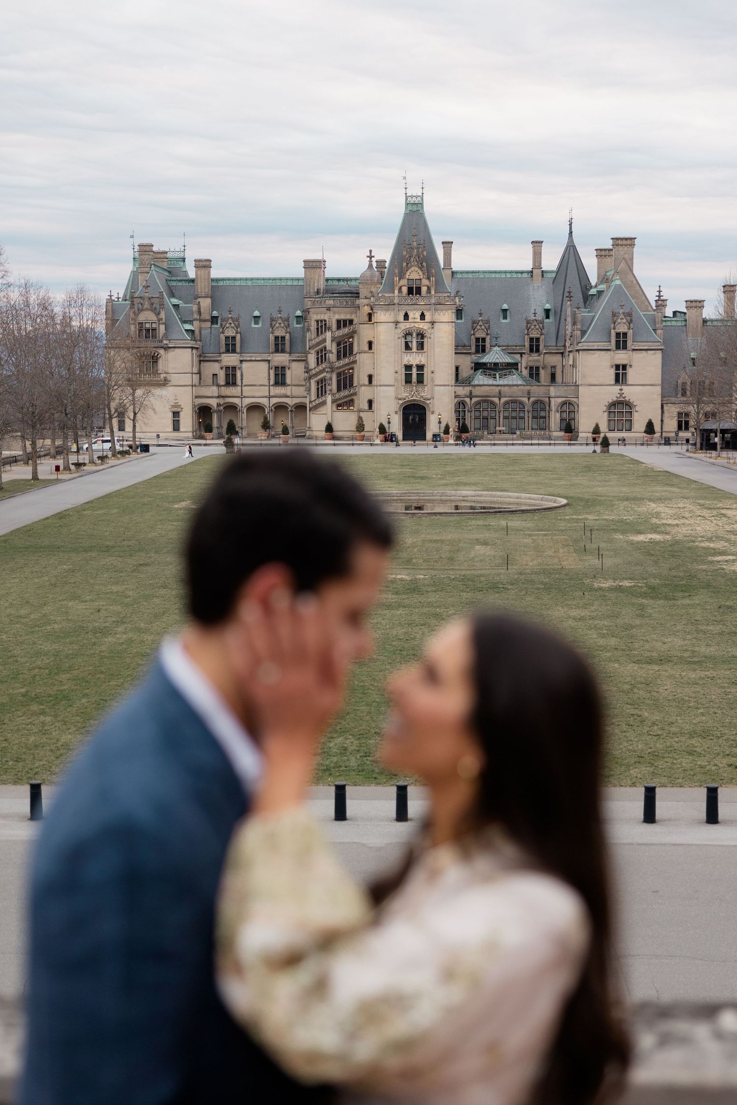 A couple poses romantically in front of the historic Biltmore Estate mansion in Asheville, North Carolina on a cloudy day.