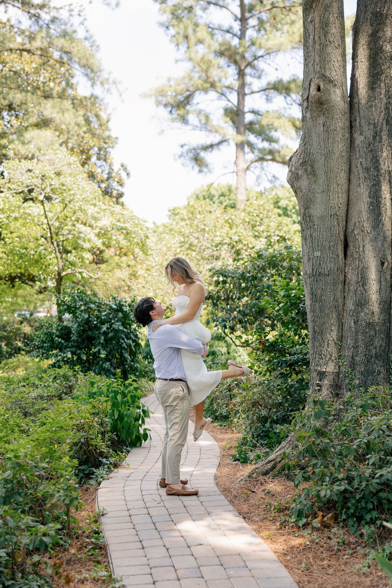 Man lifting woman in white dress on a brick garden path surrounded by tall trees and greenery.