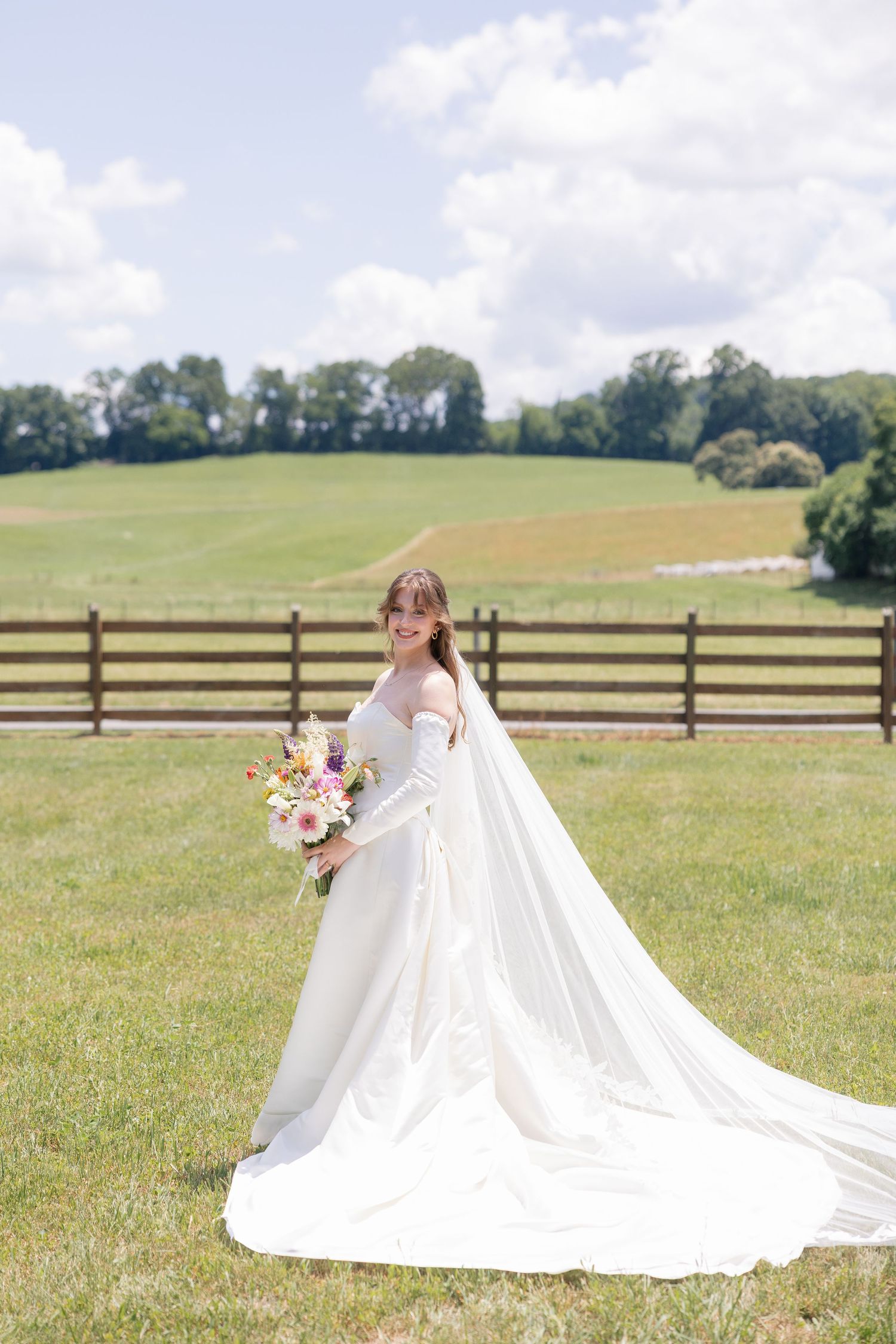 Smiling bride in off-shoulder gown with long train holds colorful bouquet by rustic fence.