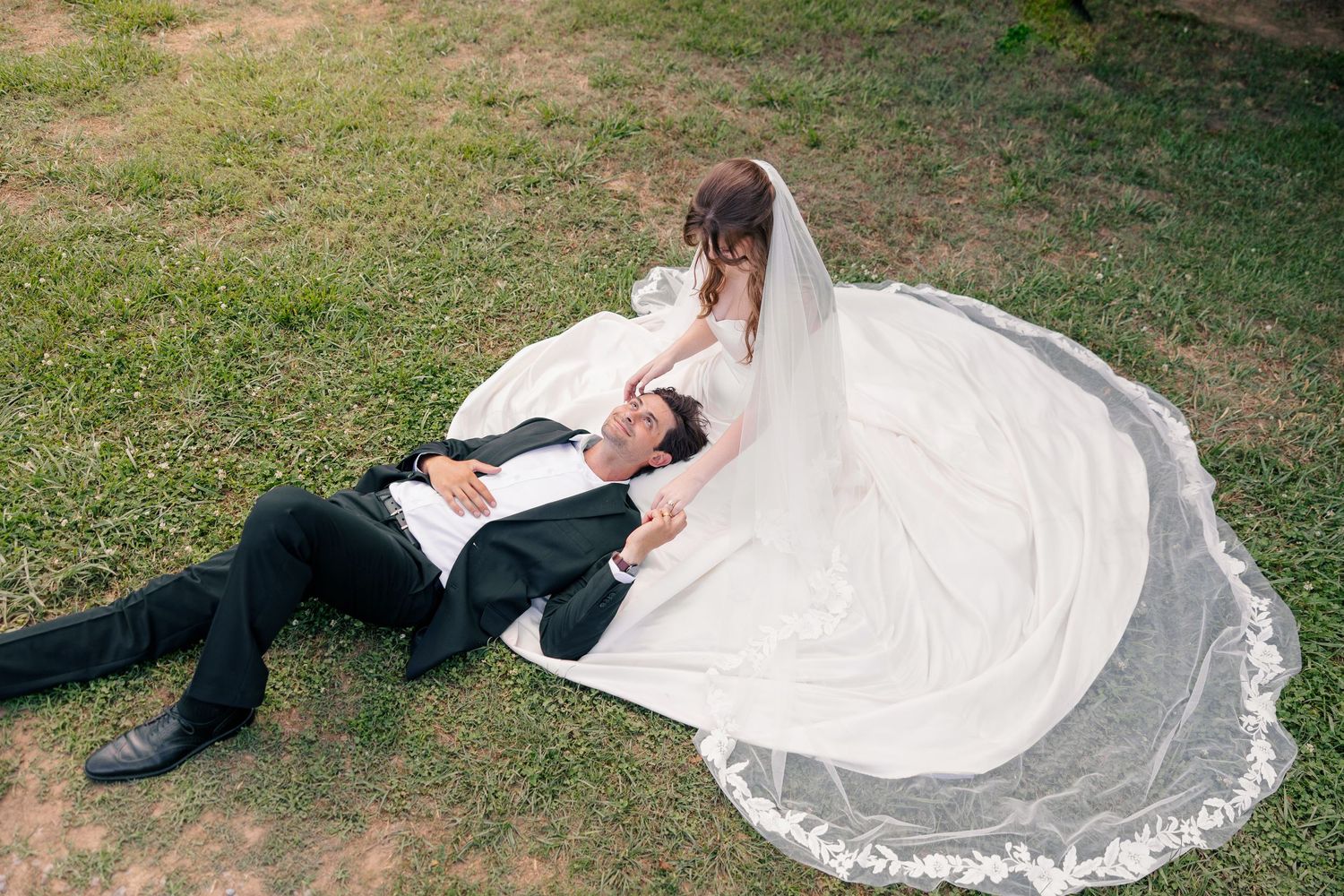 Bride in white gown and veil sits beside groom in black suit lying on green grass in romantic wedding photo.
