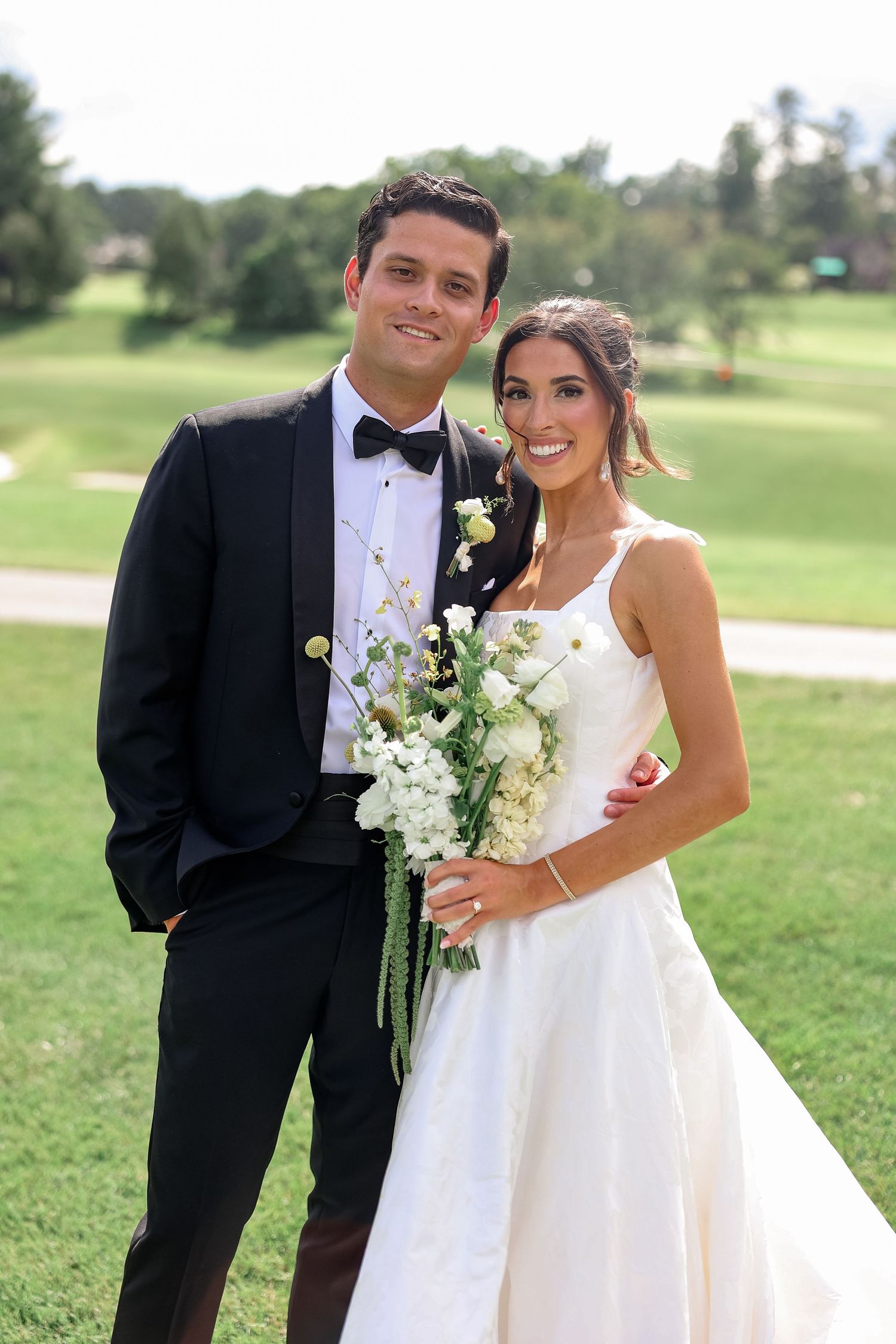 Smiling bride and groom pose together outdoors on a golf course, bride holding white and green bouquet.