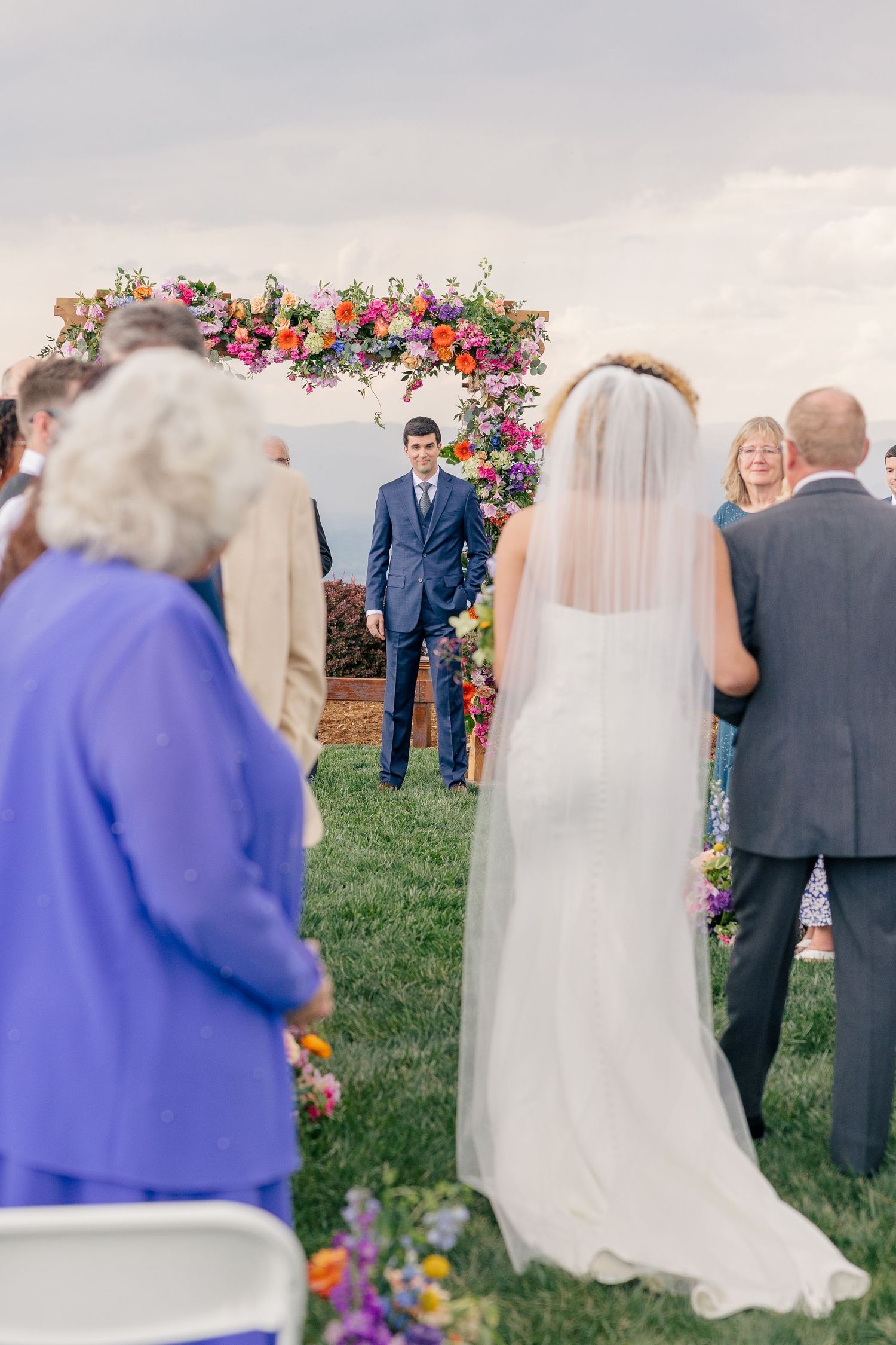 Bride walking down the aisle toward groom at colorful floral arch outdoor wedding ceremony.
