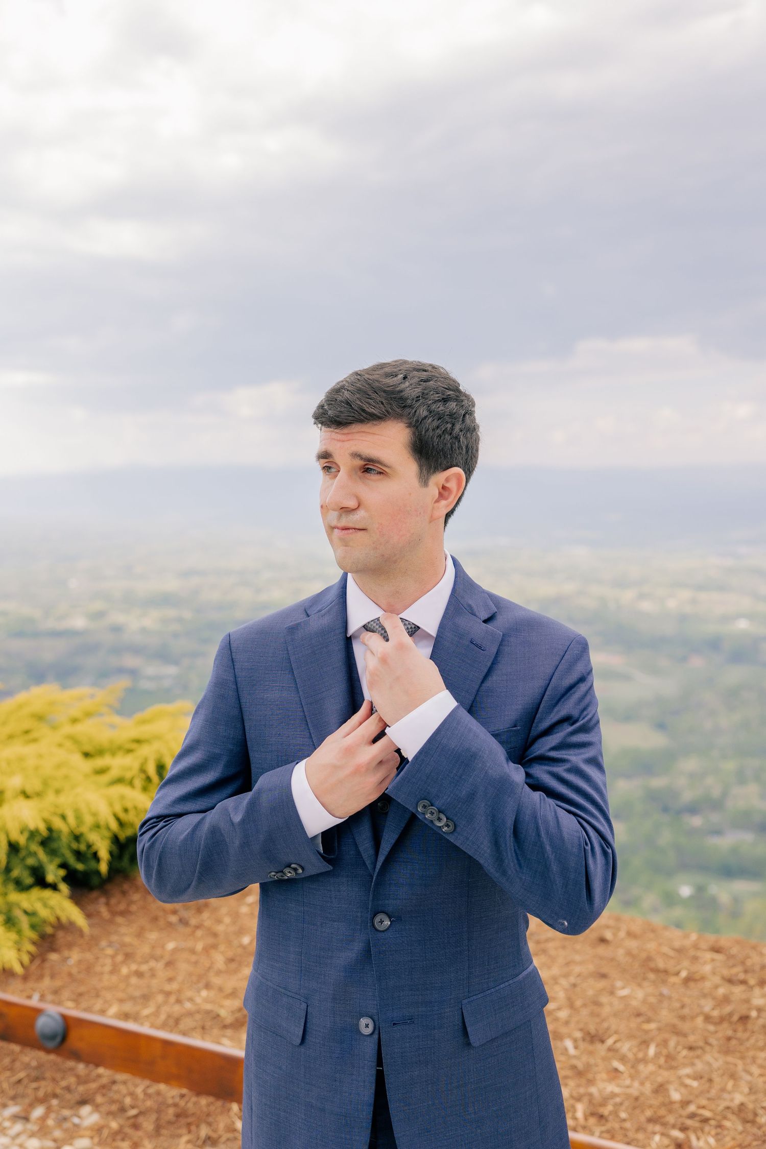 Dapper groom in navy blue suit adjusting his tie outdoors with a panoramic landscape backdrop.