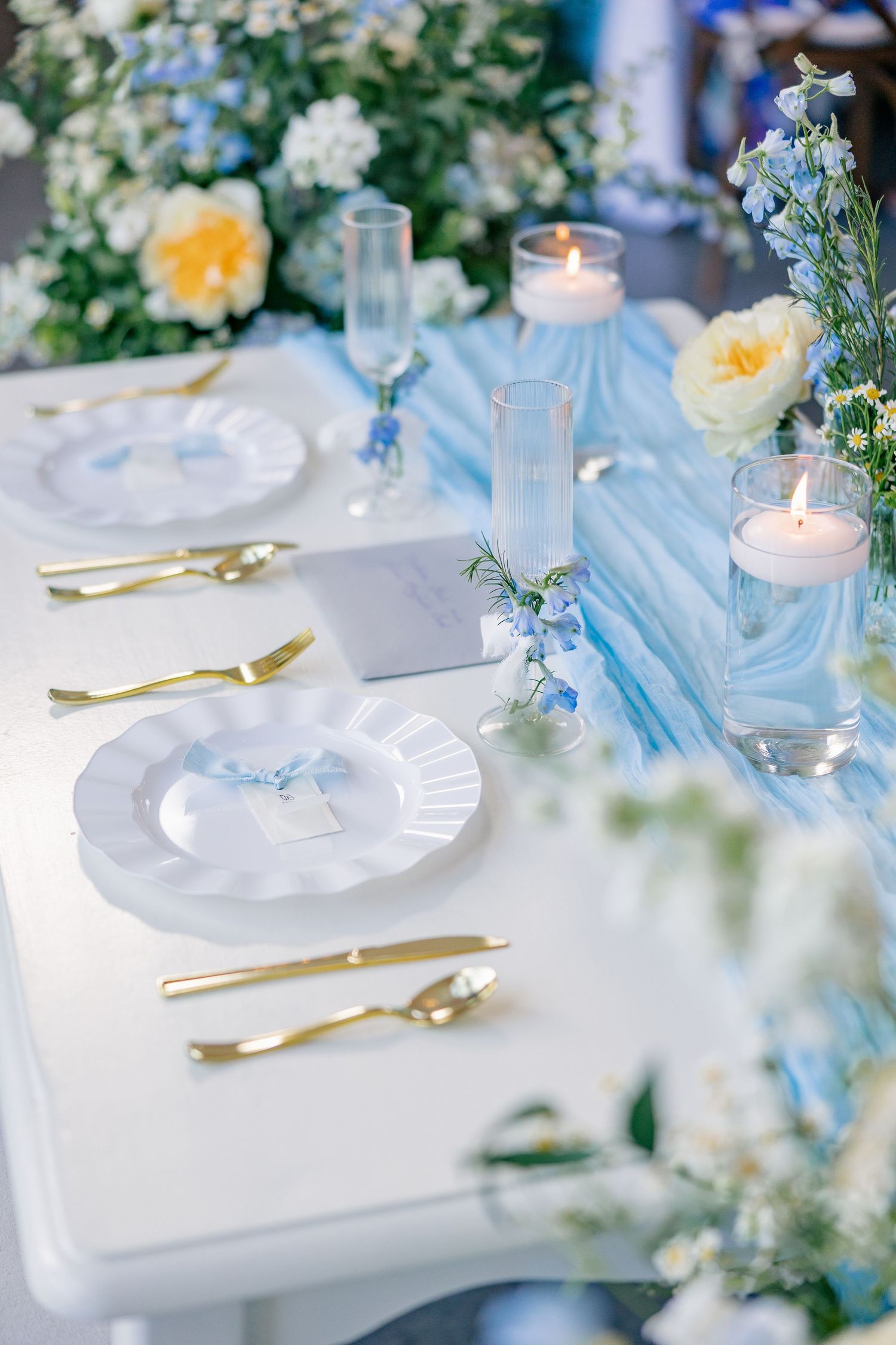 Elegant wedding table setting with gold cutlery, white plates, blue runner, candles, and yellow and blue flowers.