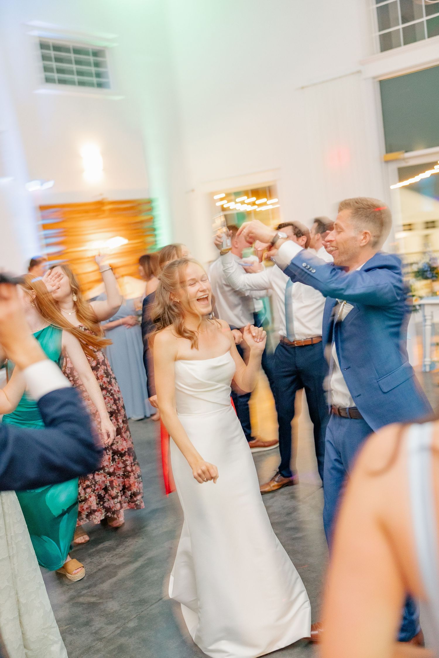 A laughing bride in white dances joyfully with guests during a lively indoor wedding reception.