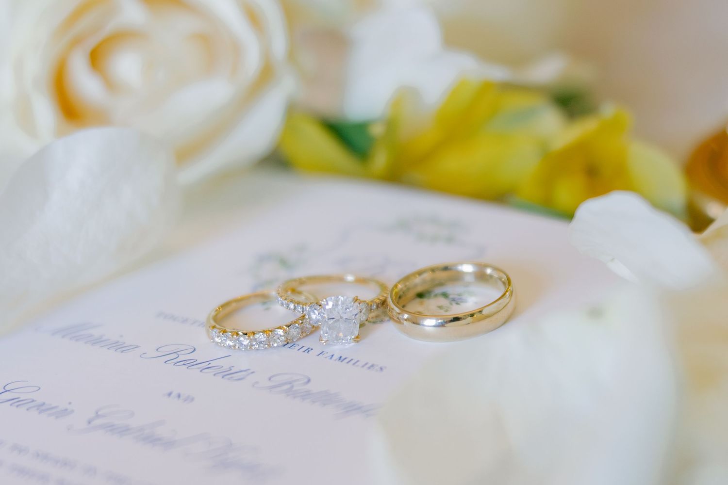 Three elegant wedding rings, including a diamond engagement ring, rest on a wedding program surrounded by white and yellow roses.