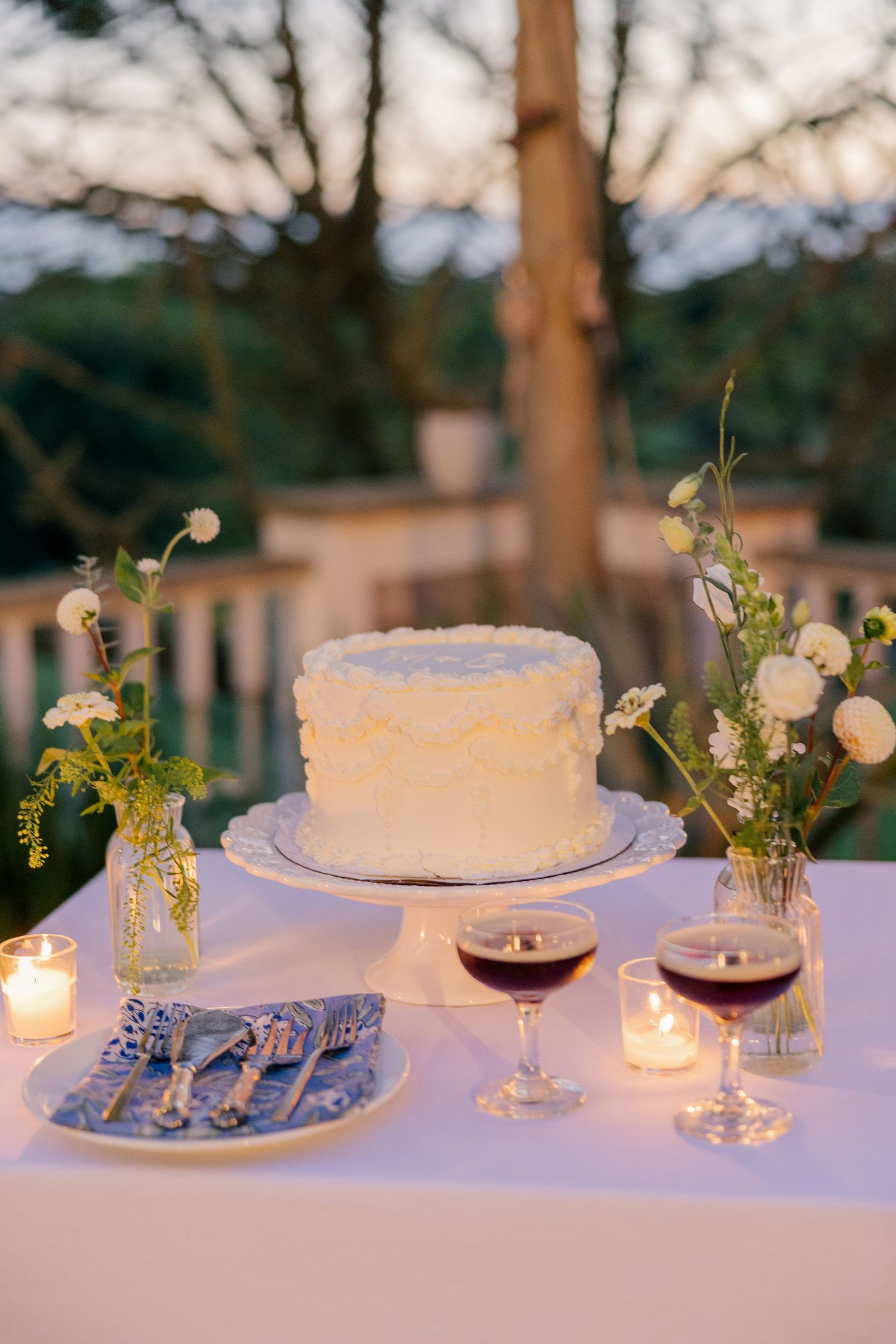 Single-tier white wedding cake on a pedestal surrounded by candles, wine glasses, and floral arrangements at dusk.