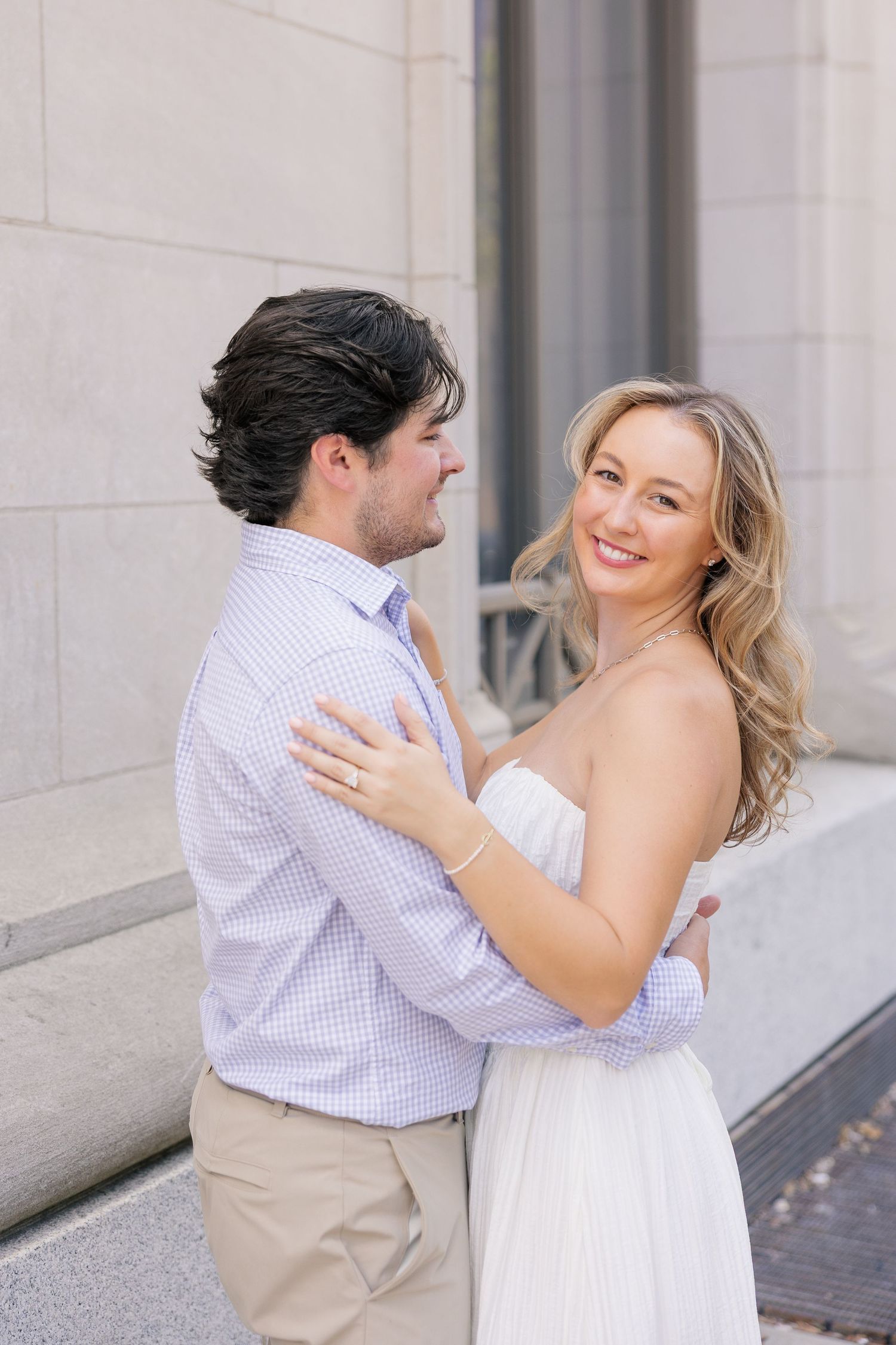 Couple sharing a romantic moment by stone pillars, woman in white strapless dress smiling at camera during engagement shoot.