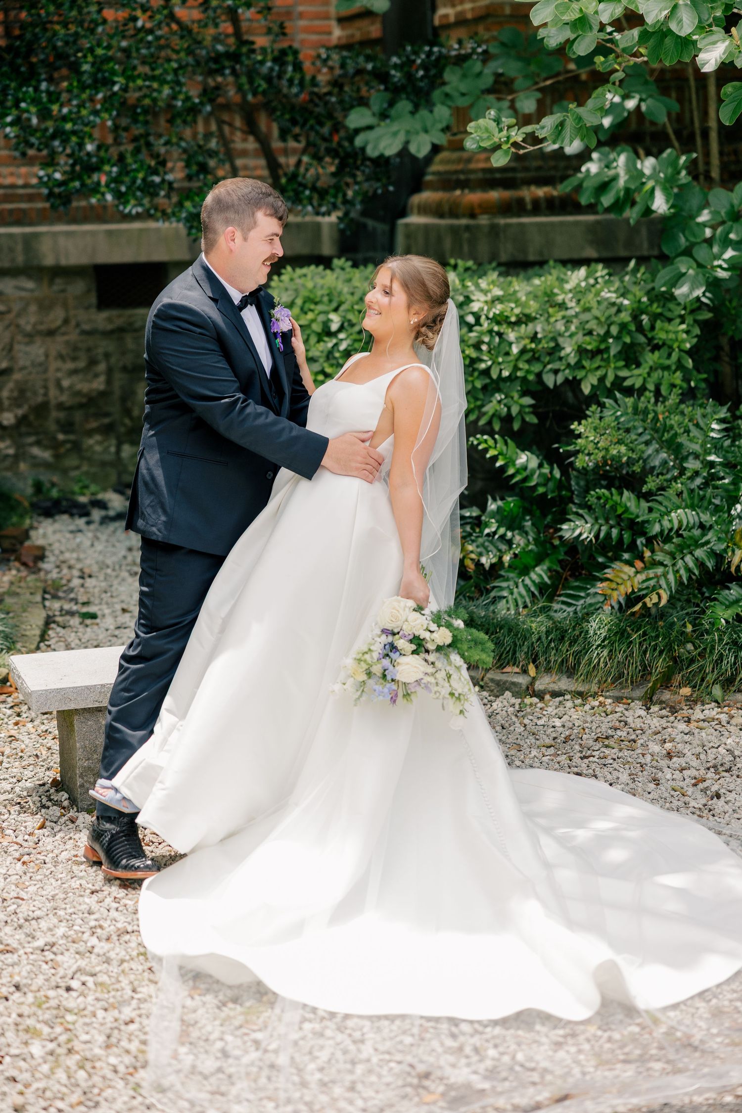 Groom embraces bride holding bouquet in lush garden setting on wedding day.