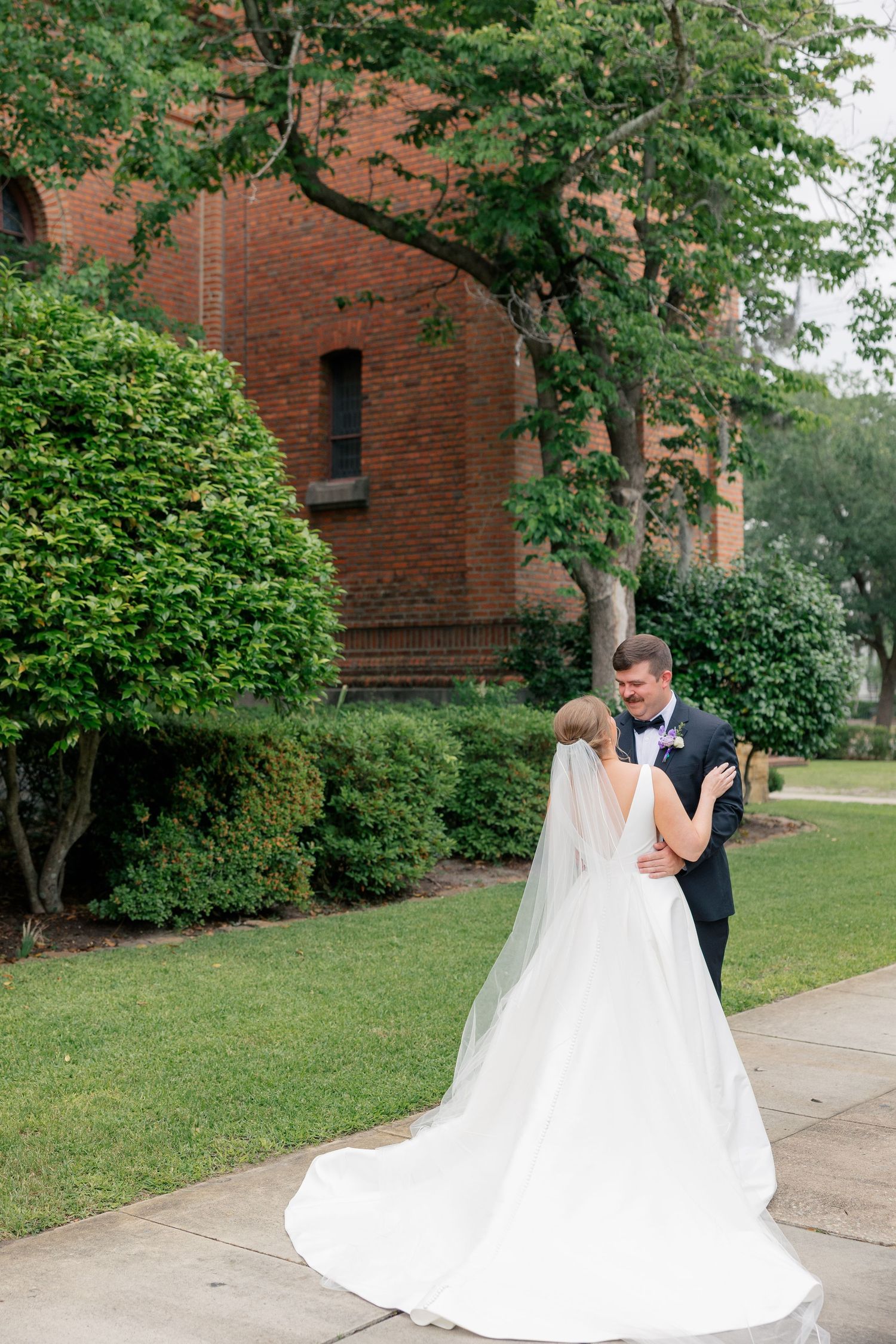 Bride in elegant white gown with veil shares romantic first look moment with groom on church grounds.