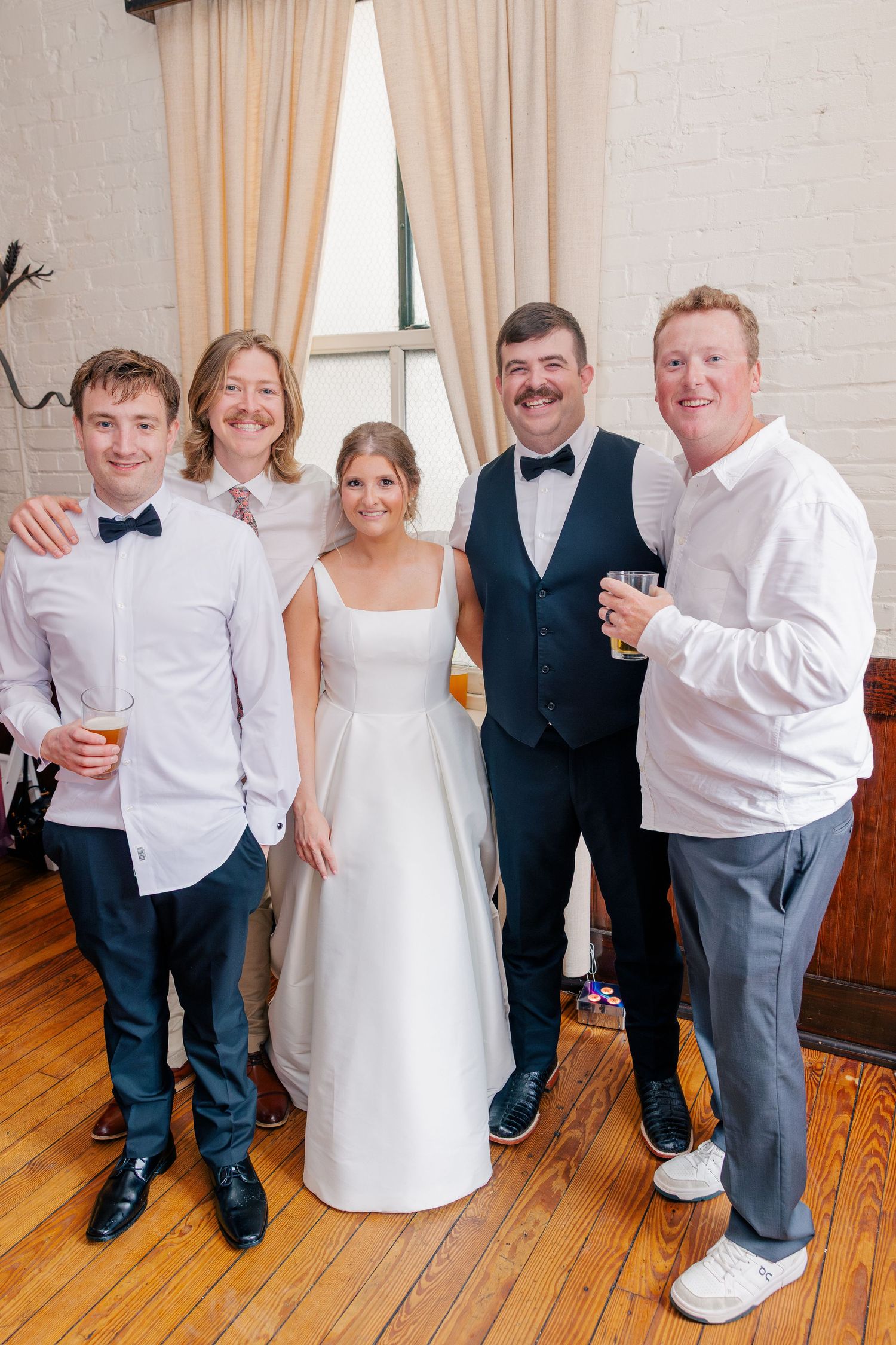 Wedding party of five posing indoors, bride in white gown flanked by groom in navy vest and bow tie with three guests.