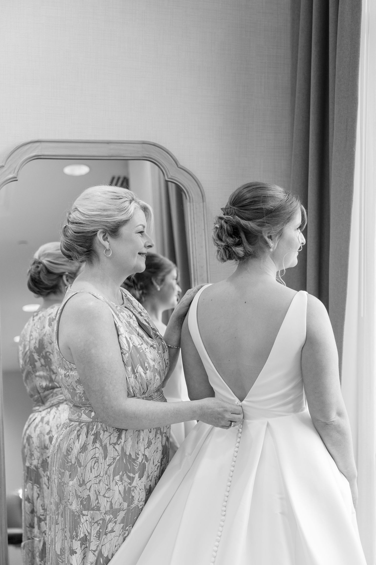 Mother helps bride button her elegant white wedding gown in a softly lit room before the ceremony.
