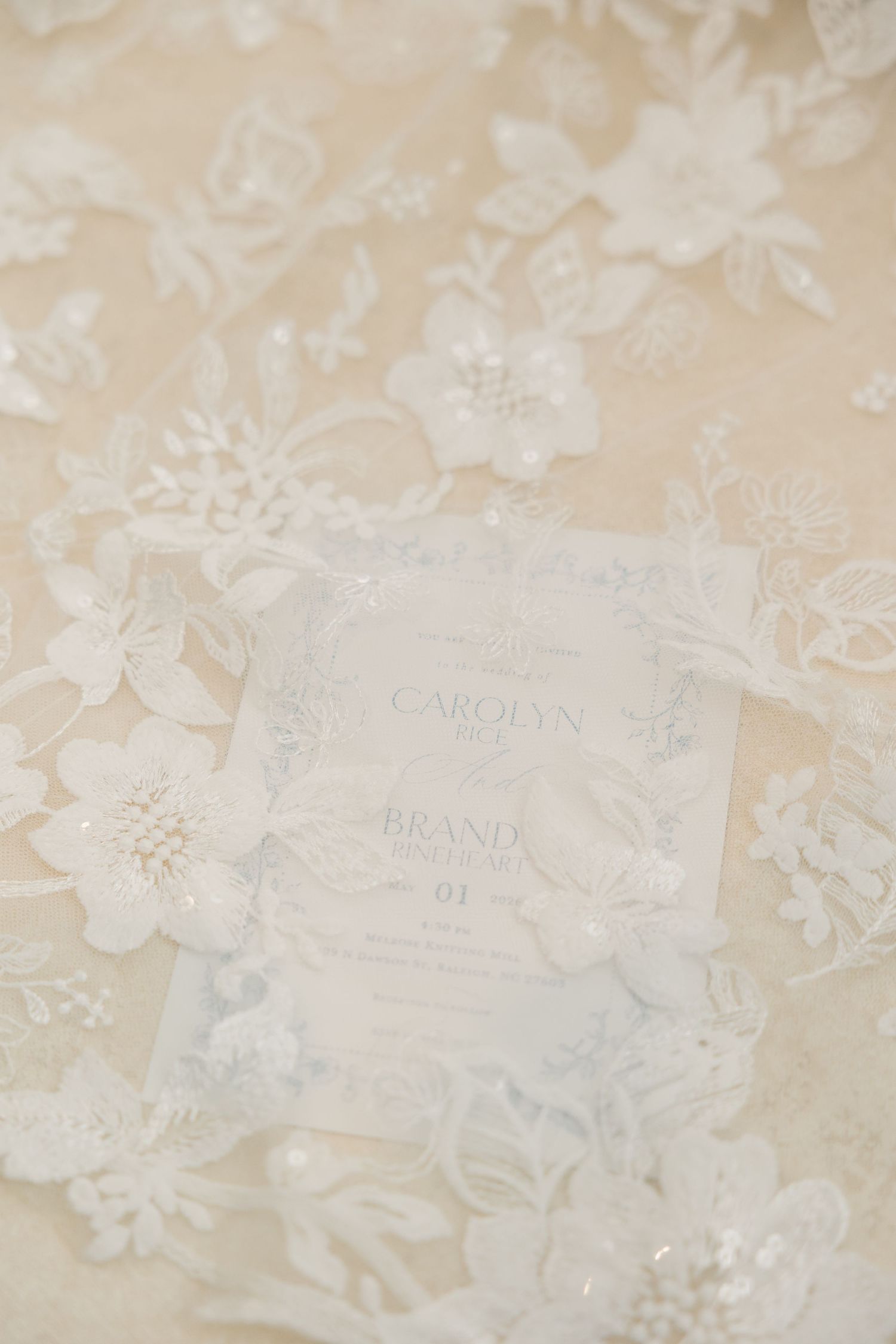 Elegant wedding invitation for Carolyn and Brand displayed on delicate white floral lace fabric with subtle embroidery details.