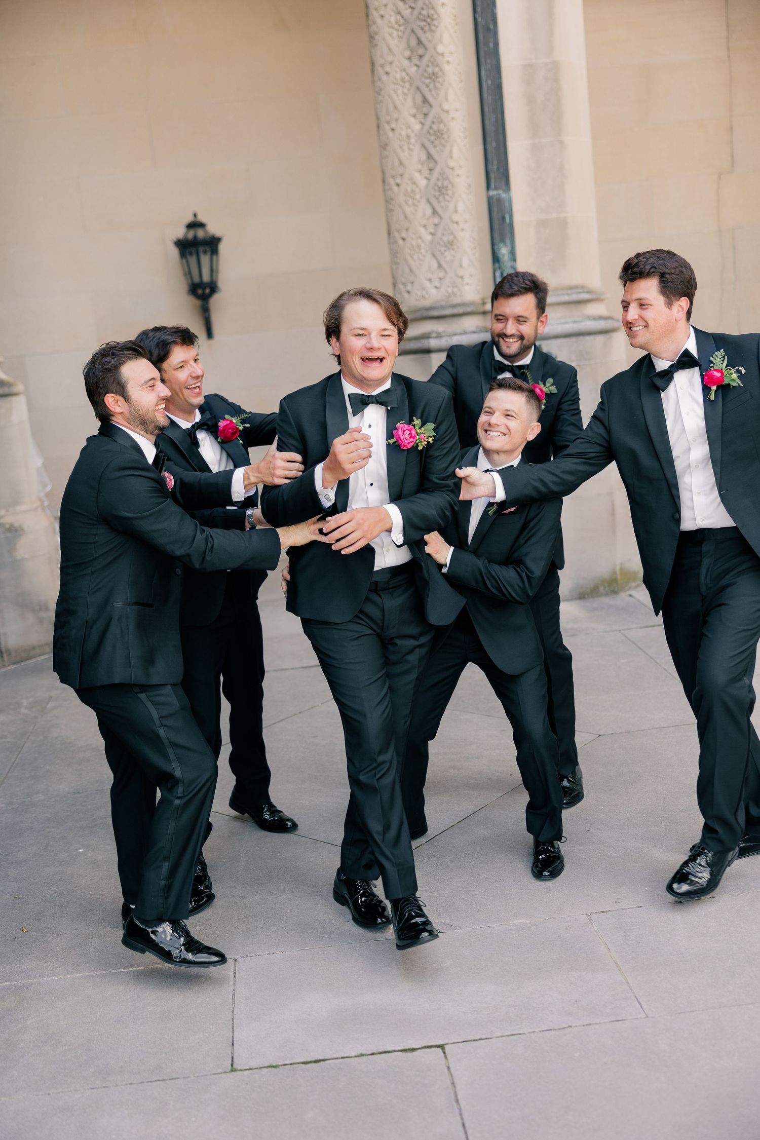 Groomsmen in black tuxedos laugh and celebrate with groom outside elegant stone building.