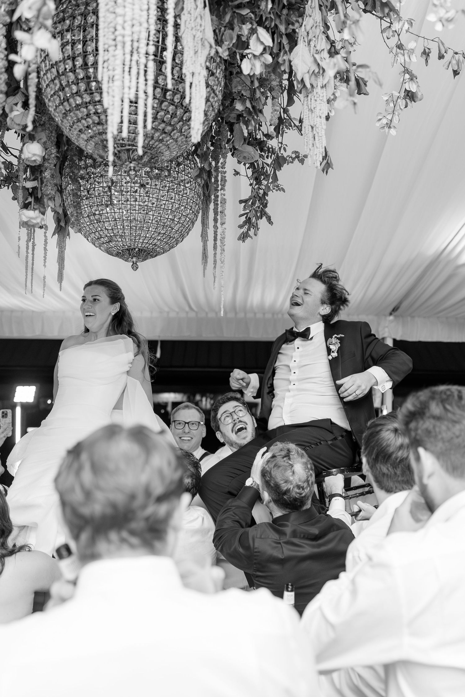 Black and white photo of bride and groom being lifted by guests during wedding hora dance.