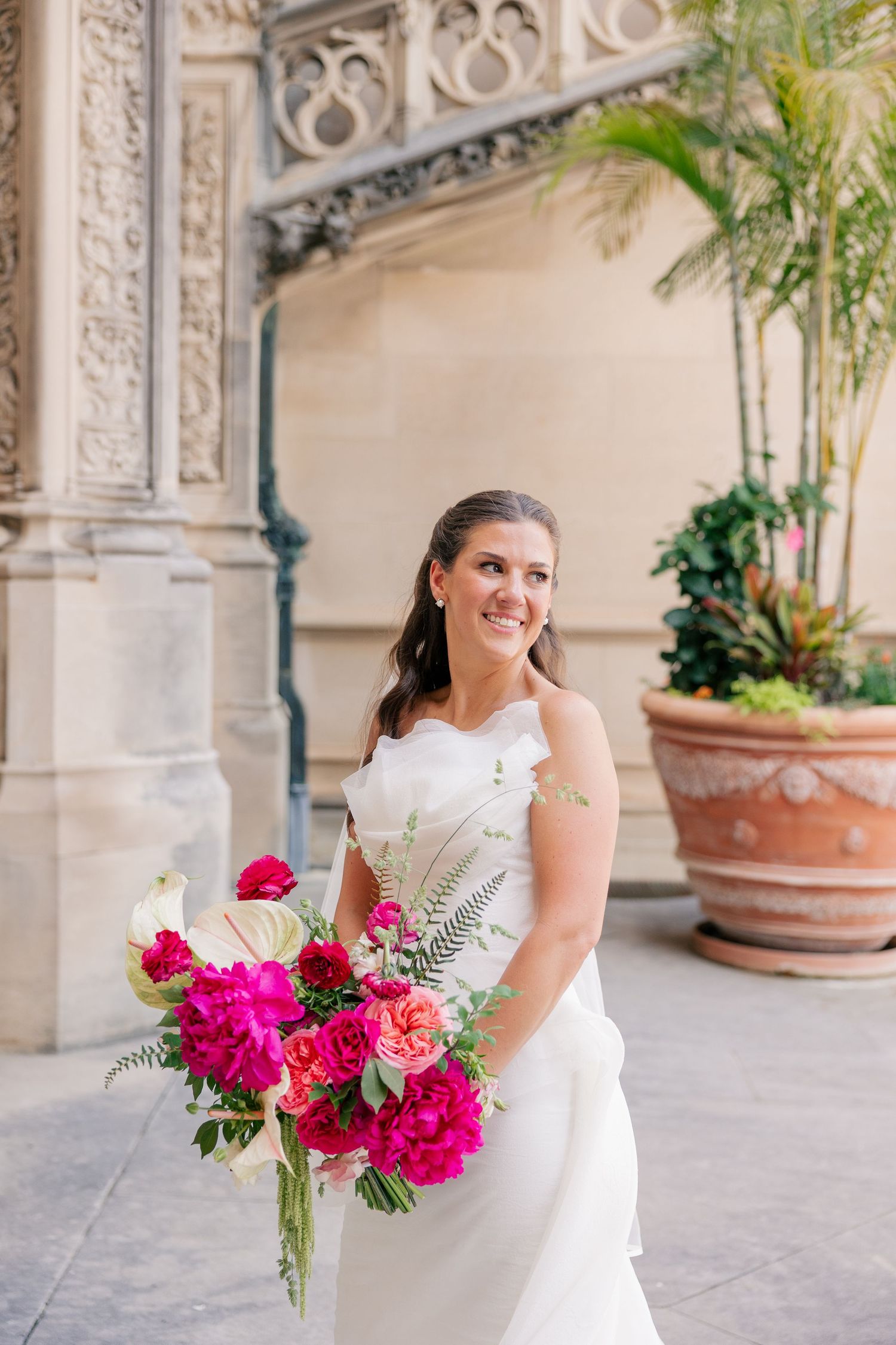 Smiling bride holds vibrant pink peony bouquet in white gown before ornate stone building.