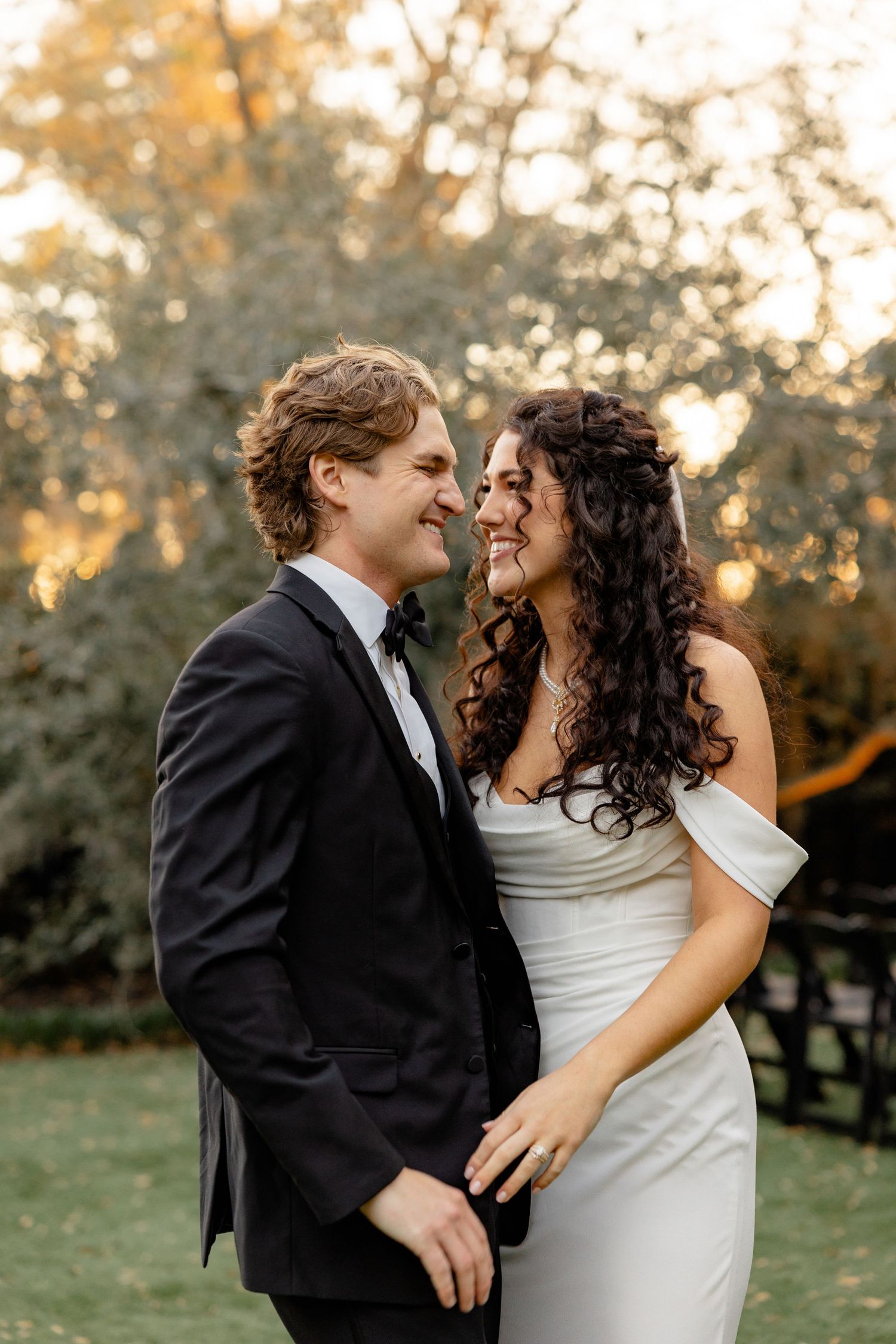 A couple shares an intimate moment at sunset in formal wedding attire on a grassy lawn.