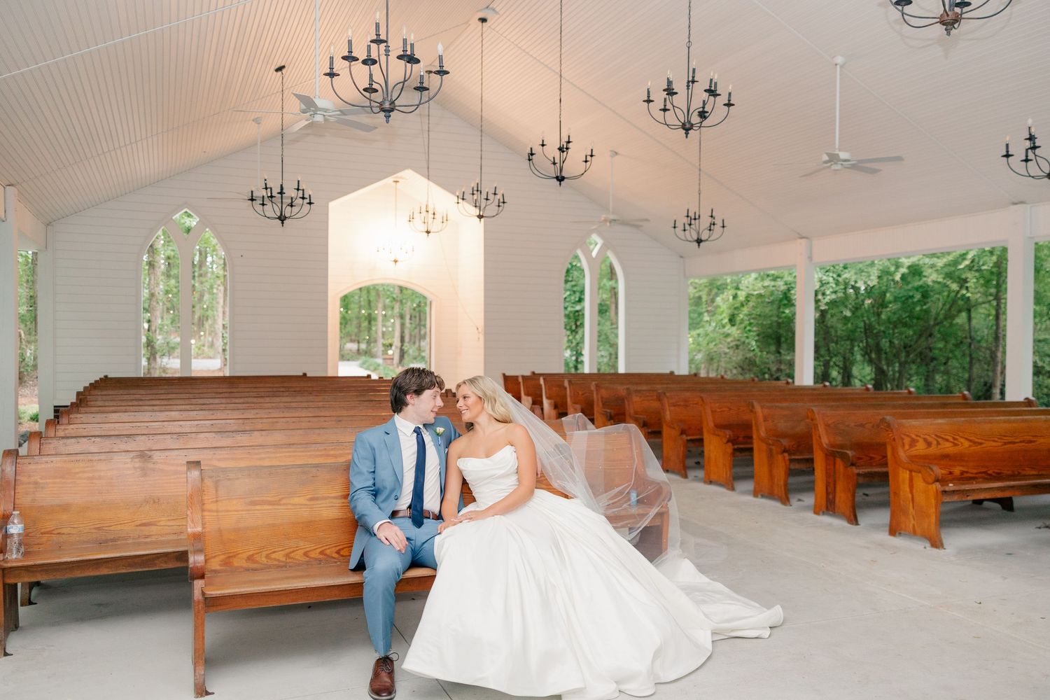 Bride and groom sit closely together on a pew inside a bright white chapel.