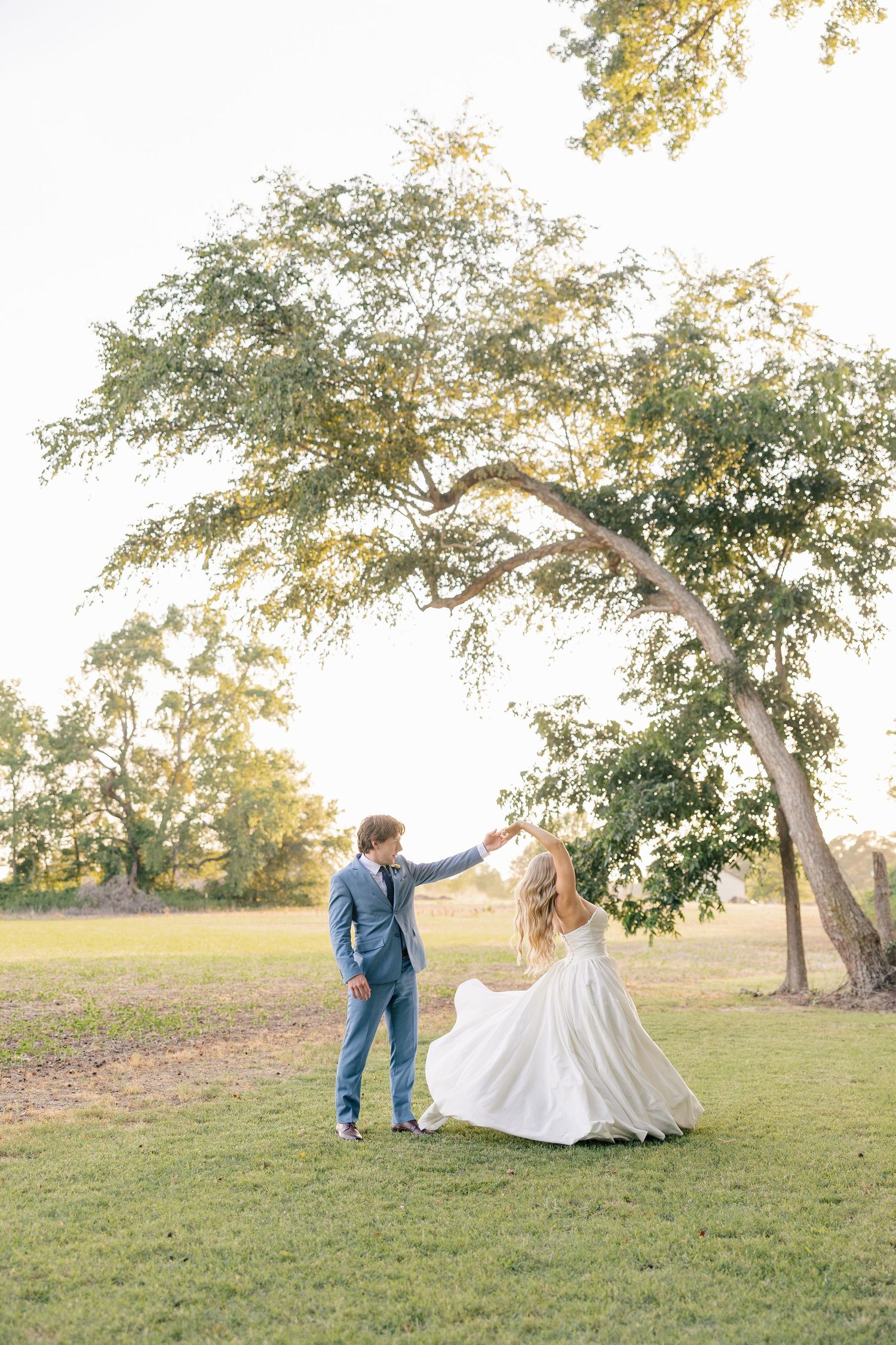 Groom spinning bride in her flowing white dress under a large tree during golden hour wedding portraits.