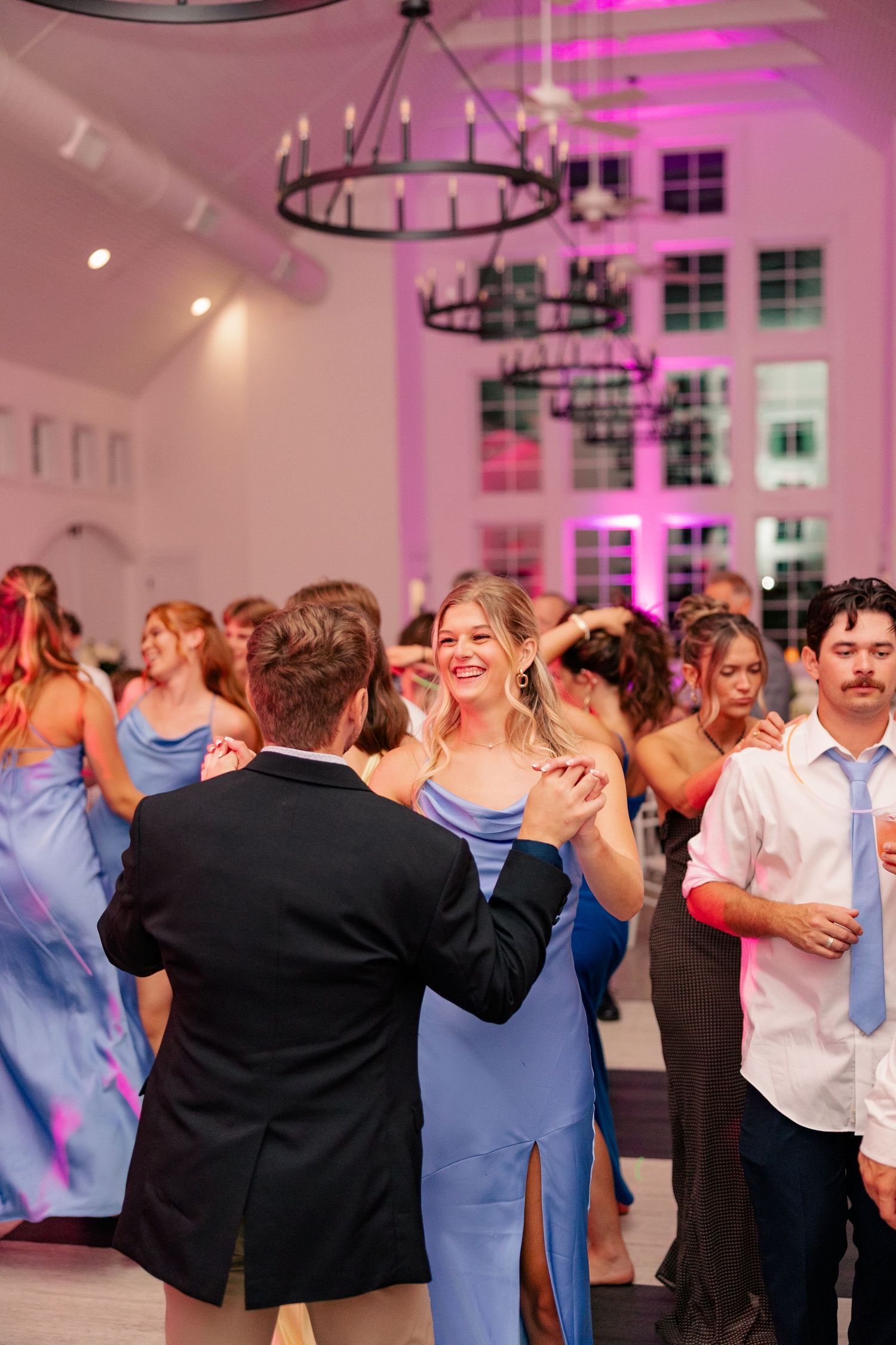 Bridesmaid in blue satin gown smiling while dancing at a pink-lit wedding reception.