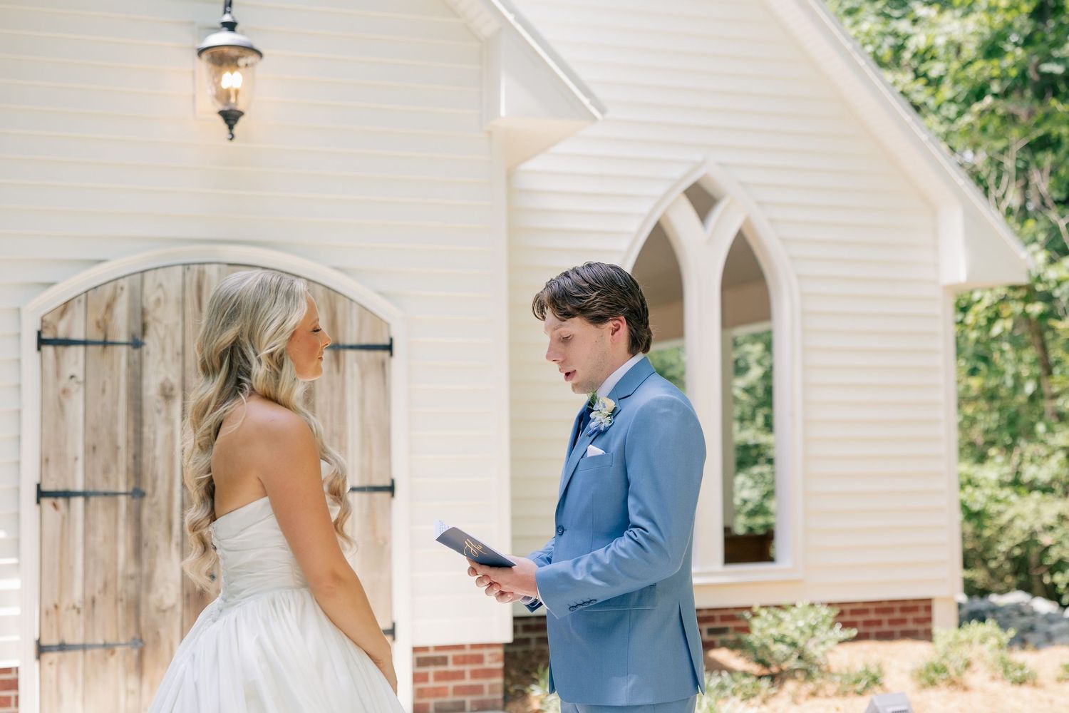 Groom in light blue suit reads vows to bride in strapless gown outside a charming white chapel.