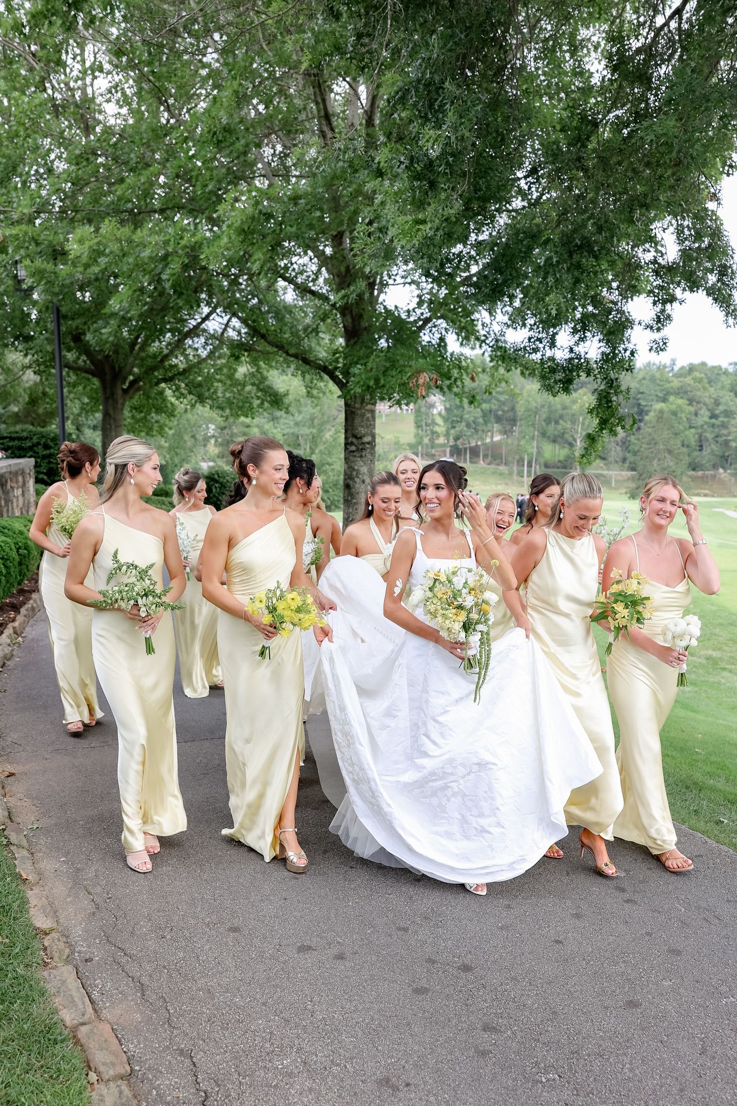 Bride in white gown walks with bridesmaids in yellow dresses, laughing outdoors near trees on a wedding day.