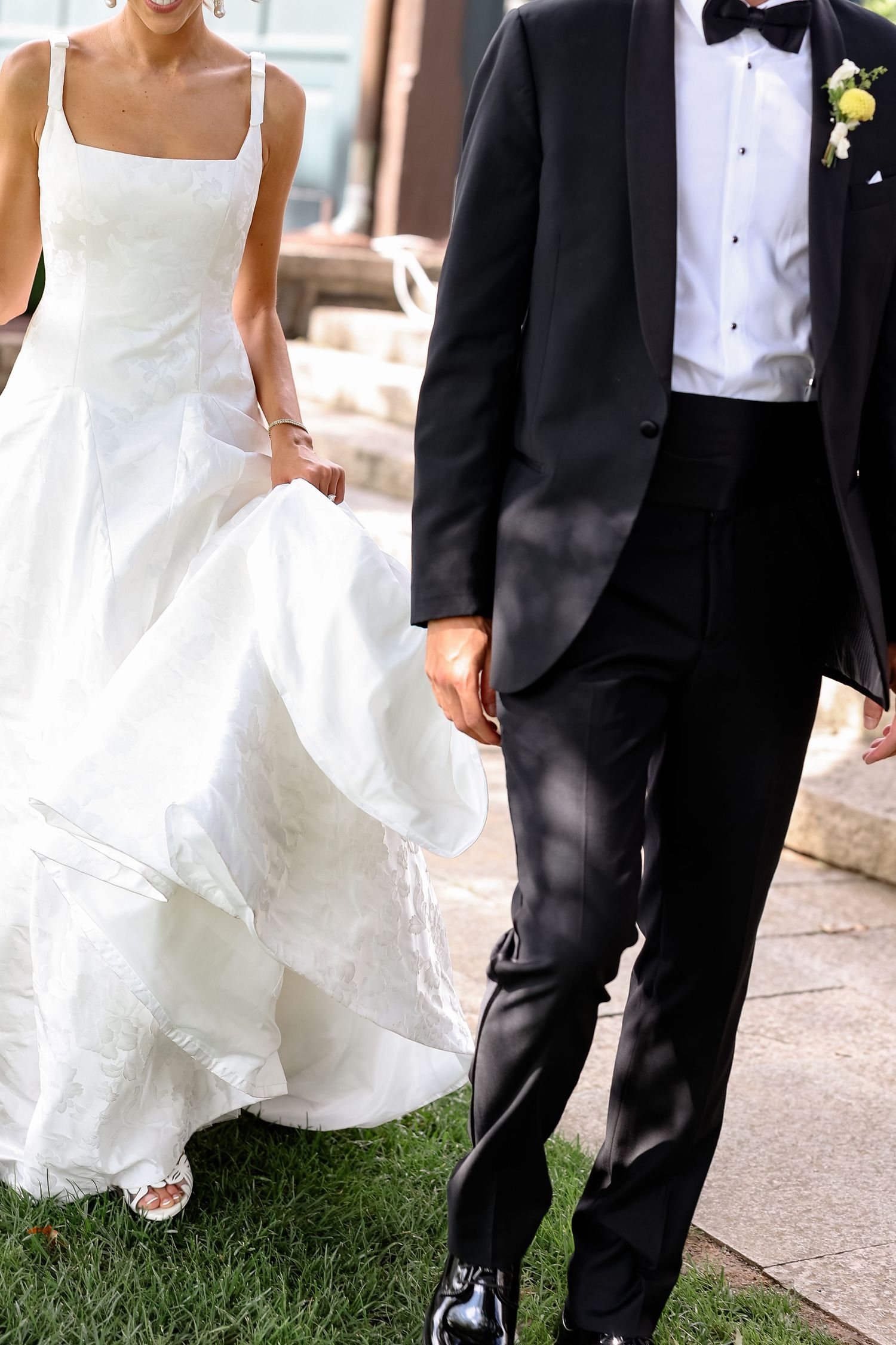 Bride in white ball gown and groom in black tuxedo walking hand in hand on green grass at wedding.