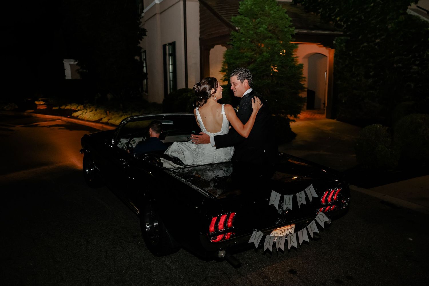 Bride and groom share a romantic embrace beside a classic black car at night during their wedding celebration.
