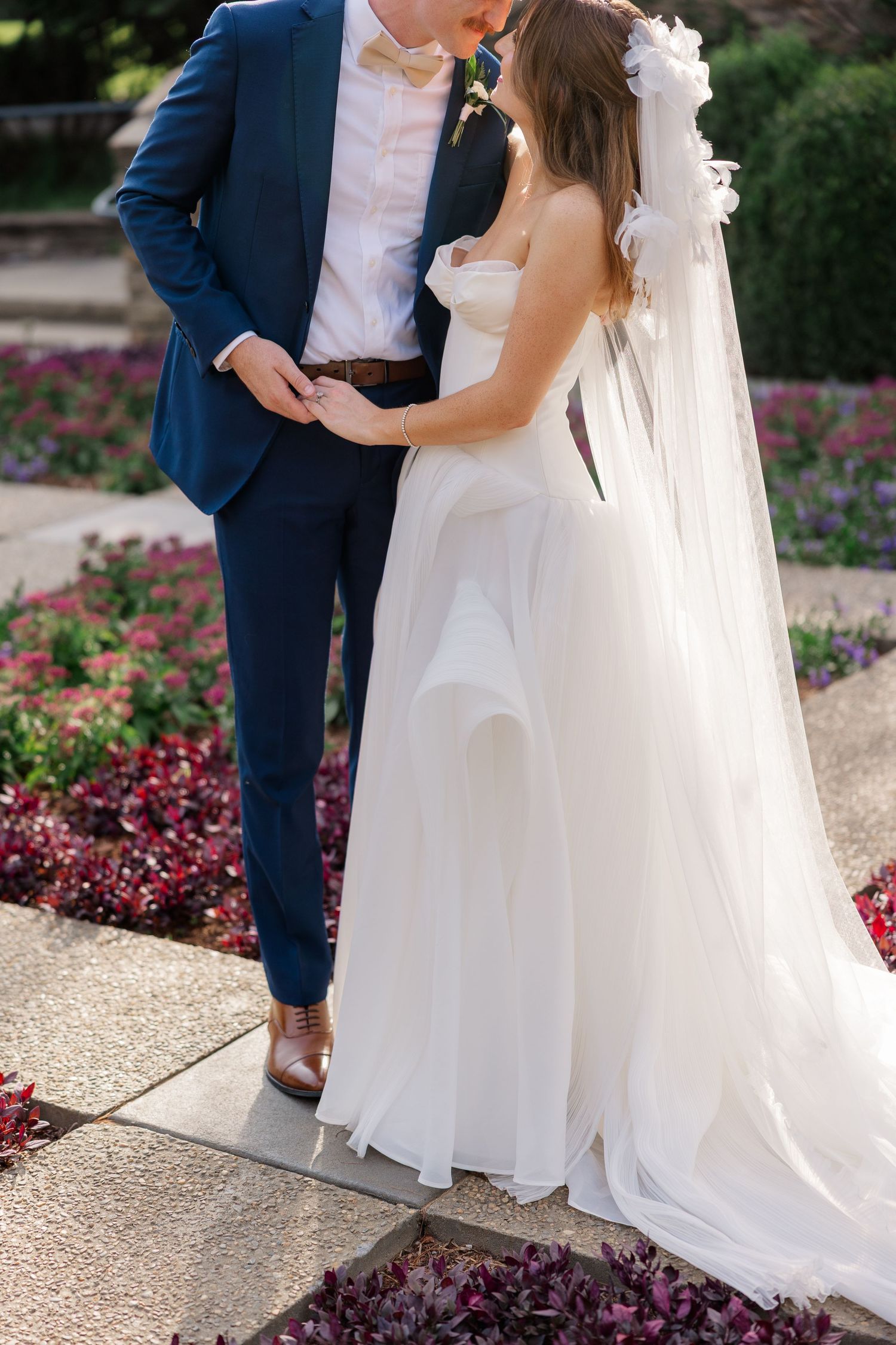 A couple shares an intimate moment in a garden setting with burgundy flowers and white wedding attire.