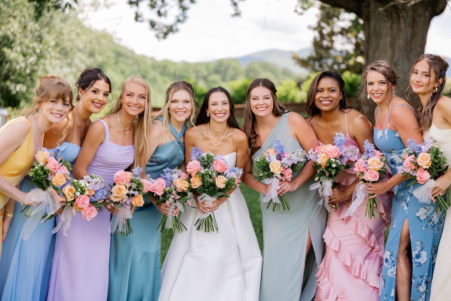 Bridesmaids in pastel blue and pink dresses hold colorful bouquets while standing together outdoors.