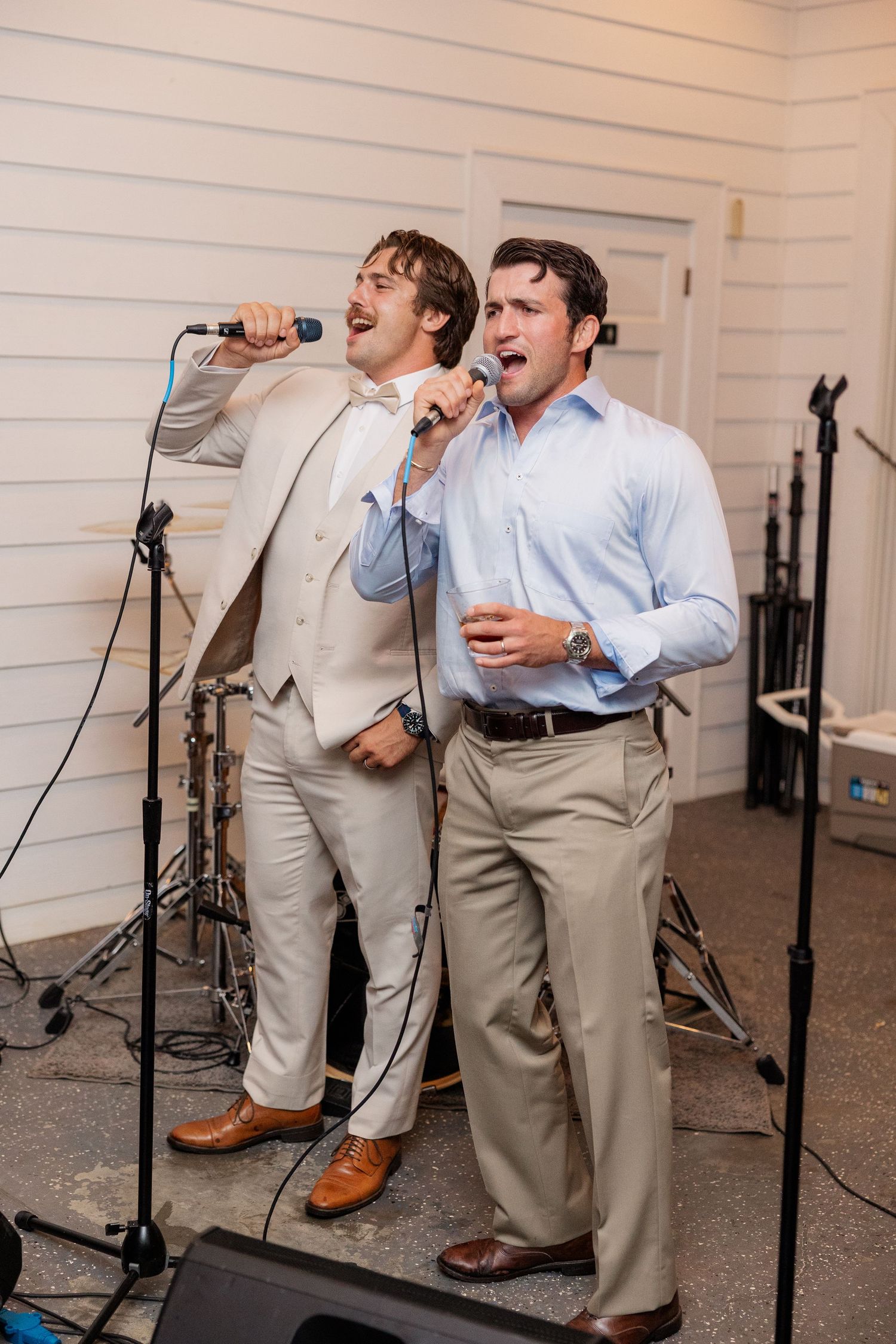 Groomsmen in light colored suits singing and performing with microphones at an indoor wedding reception.
