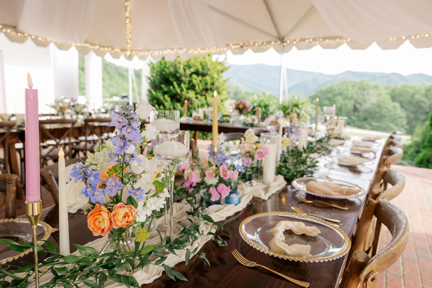 Wedding reception table settings with elegant floral arrangements and string lights under a white tent.