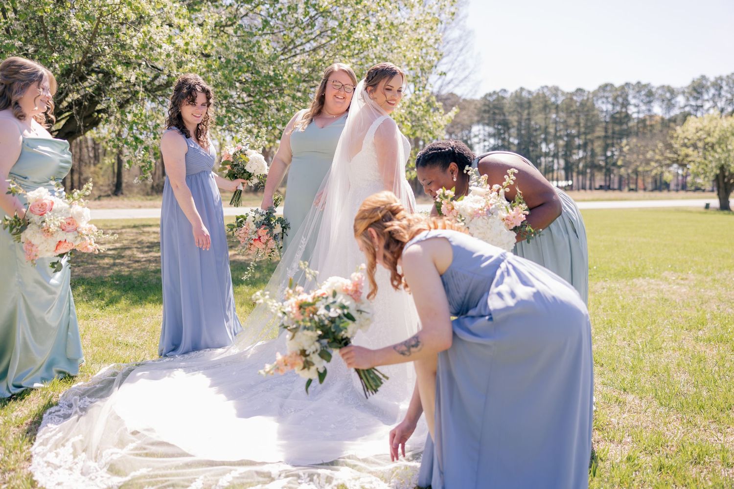 Bridesmaids in light blue dresses gather together with bouquets in a sunny garden setting.