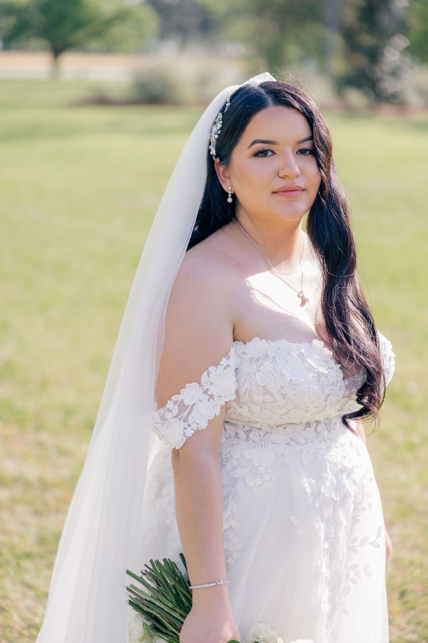 Bride in an elegant off-shoulder lace wedding gown poses outdoors holding a bouquet against a sunlit natural backdrop.