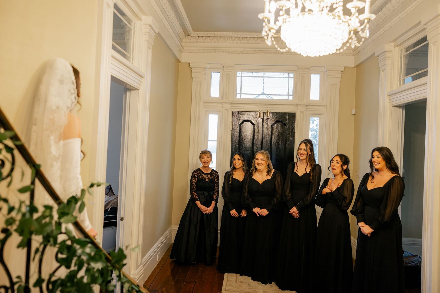 A row of bridesmaids in black dresses stand in an elegant hallway with a crystal chandelier.