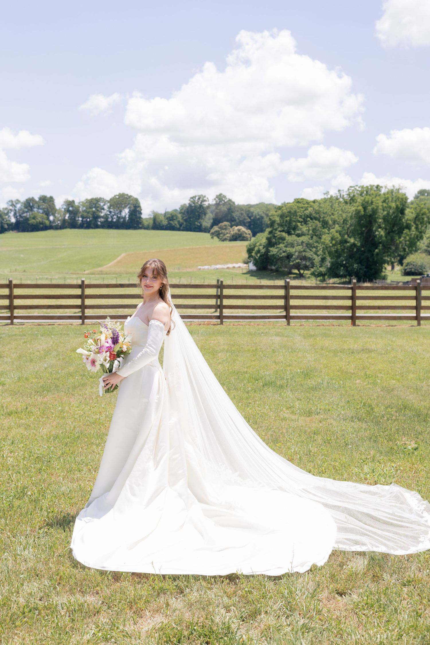 Elegant bride in off-shoulder gown with long train poses in a sunlit field holding a colorful bouquet.