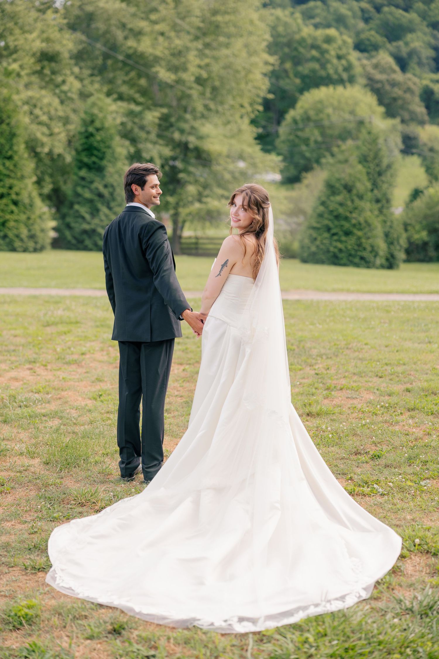 Bride and groom holding hands in green field, bride in flowing white gown with cathedral veil train.