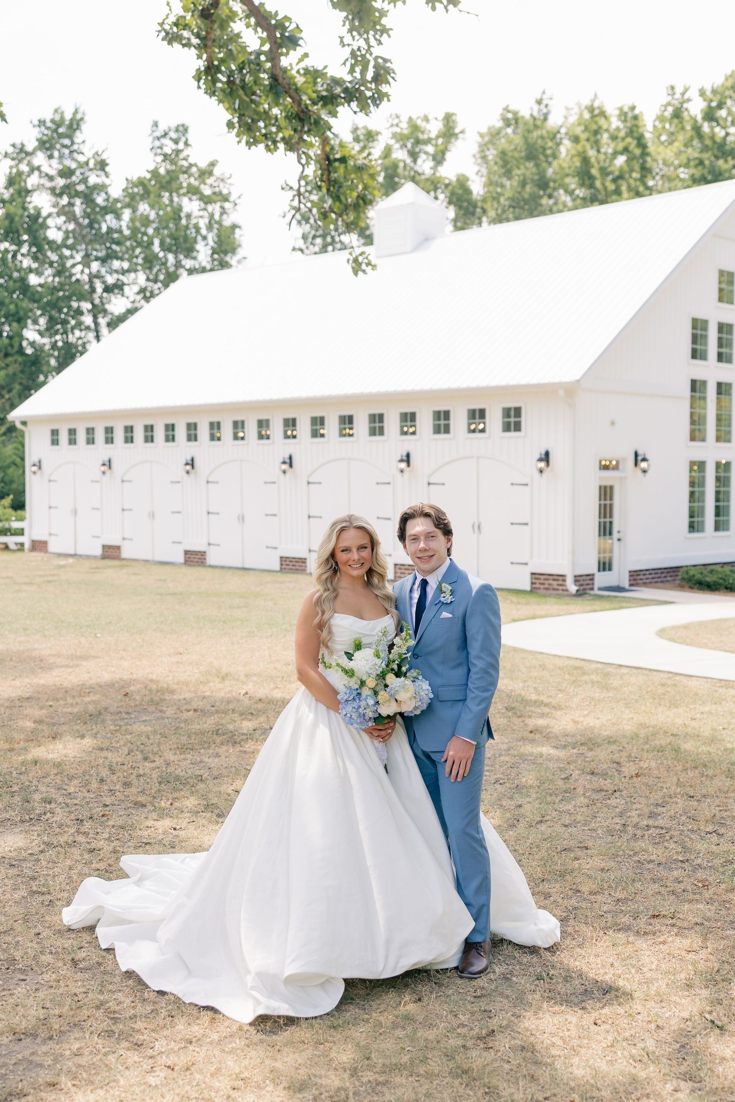 Bride in white ballgown and groom in blue suit pose together outside a white barn wedding venue.